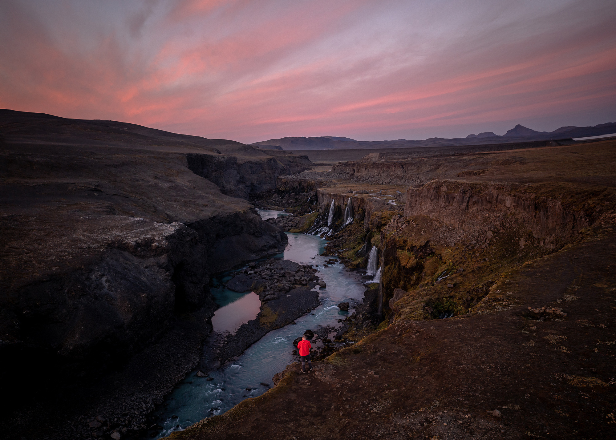 An aerial drone photo of a figure in a red coat standing at the edge of a ravine with a river at the bottom, shot at sunrise by Paul Watson with a DJI Mavic 2 Pro