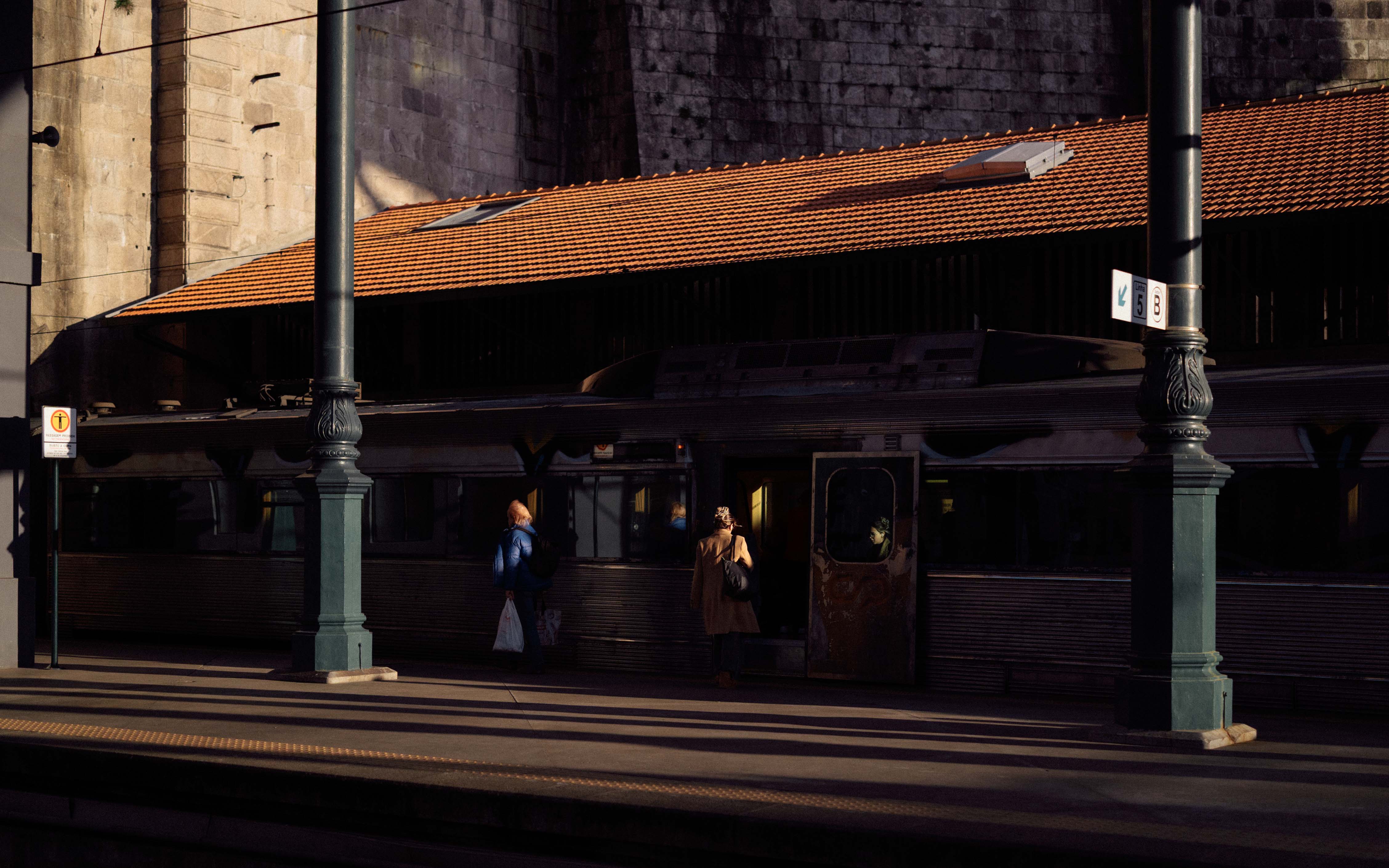Passengers boarding a train at São Bento station, with warm sunlight cutting across the platform and highlighting the train doors and stone walls behind.
