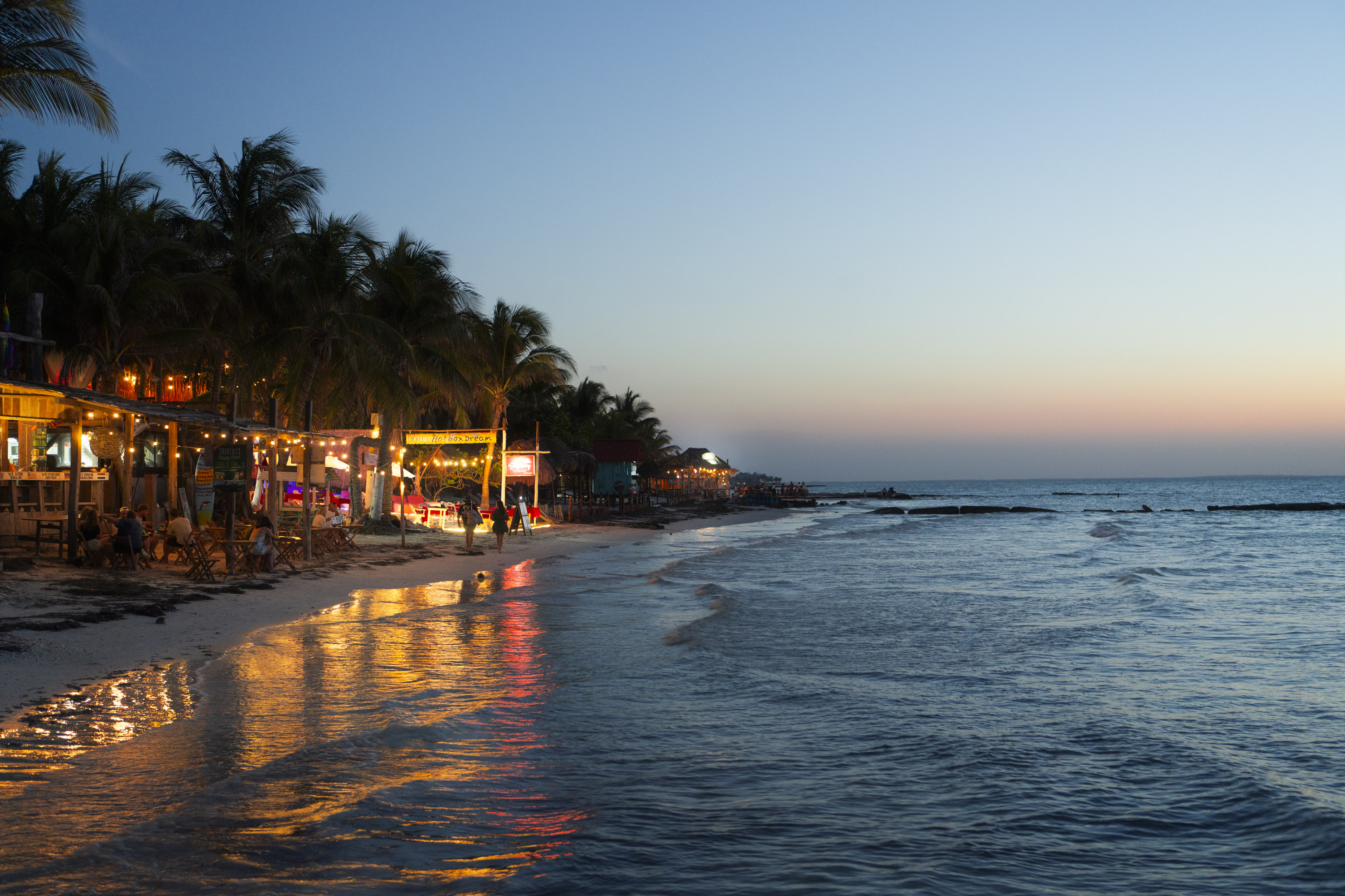A resort beach in Mexico at sunset with small bars and warm glowing lights. By Ian Howorth. 

