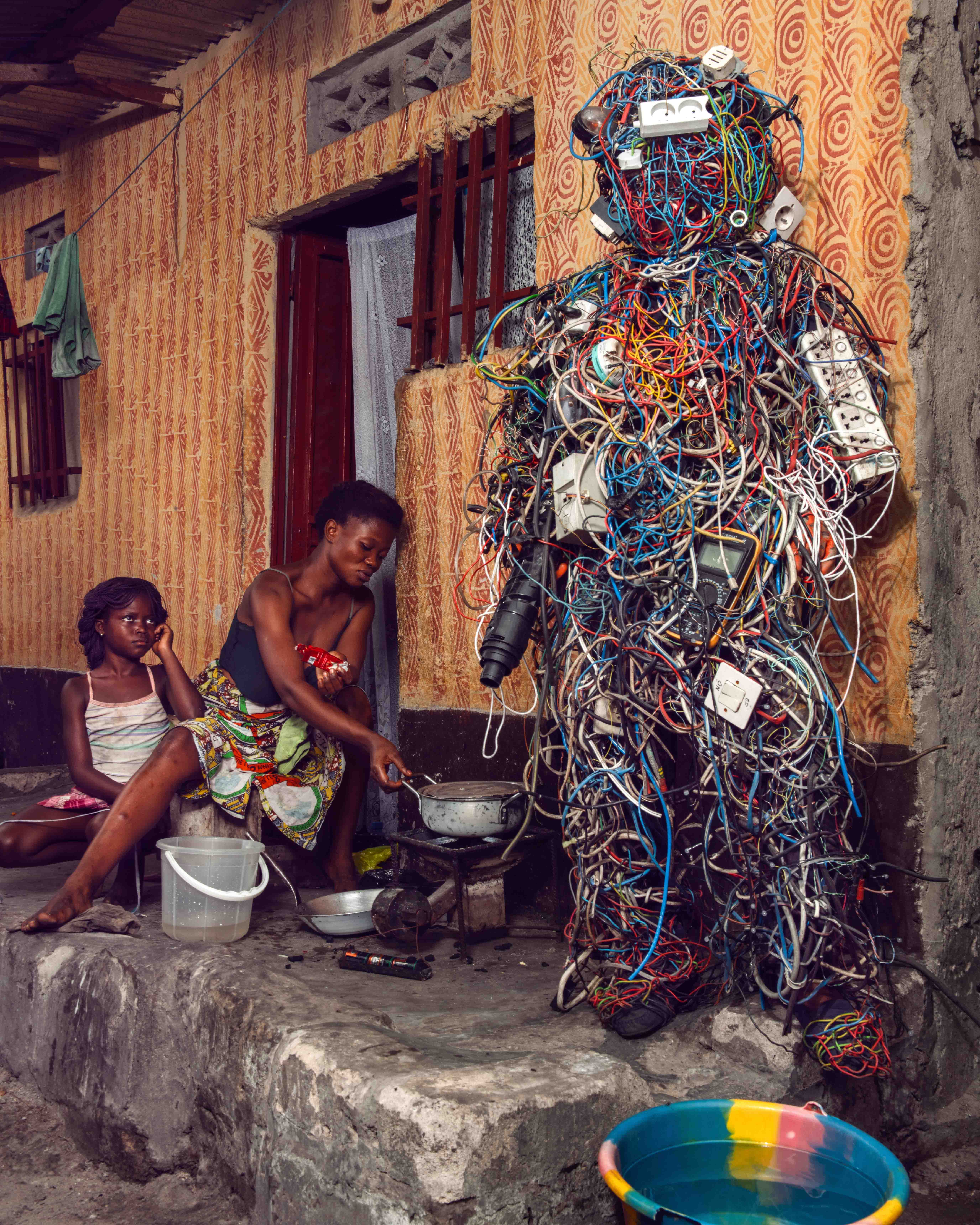 In Drähte und Stecker gehüllte Figur aus Homo Detritus, die in einem Hof in Kinshasa steht. Ein Aufeinandertreffen von Alltag und Elektroschrott.