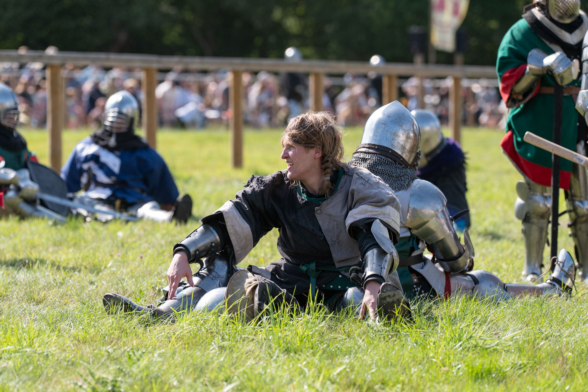 A person in a medieval costume without their helmet after battle. Their hair is messy, and they are sitting back-to-back with somebody in a knight’s costume. Other people in costume can be seen in the background, along with a crowd of spectators.