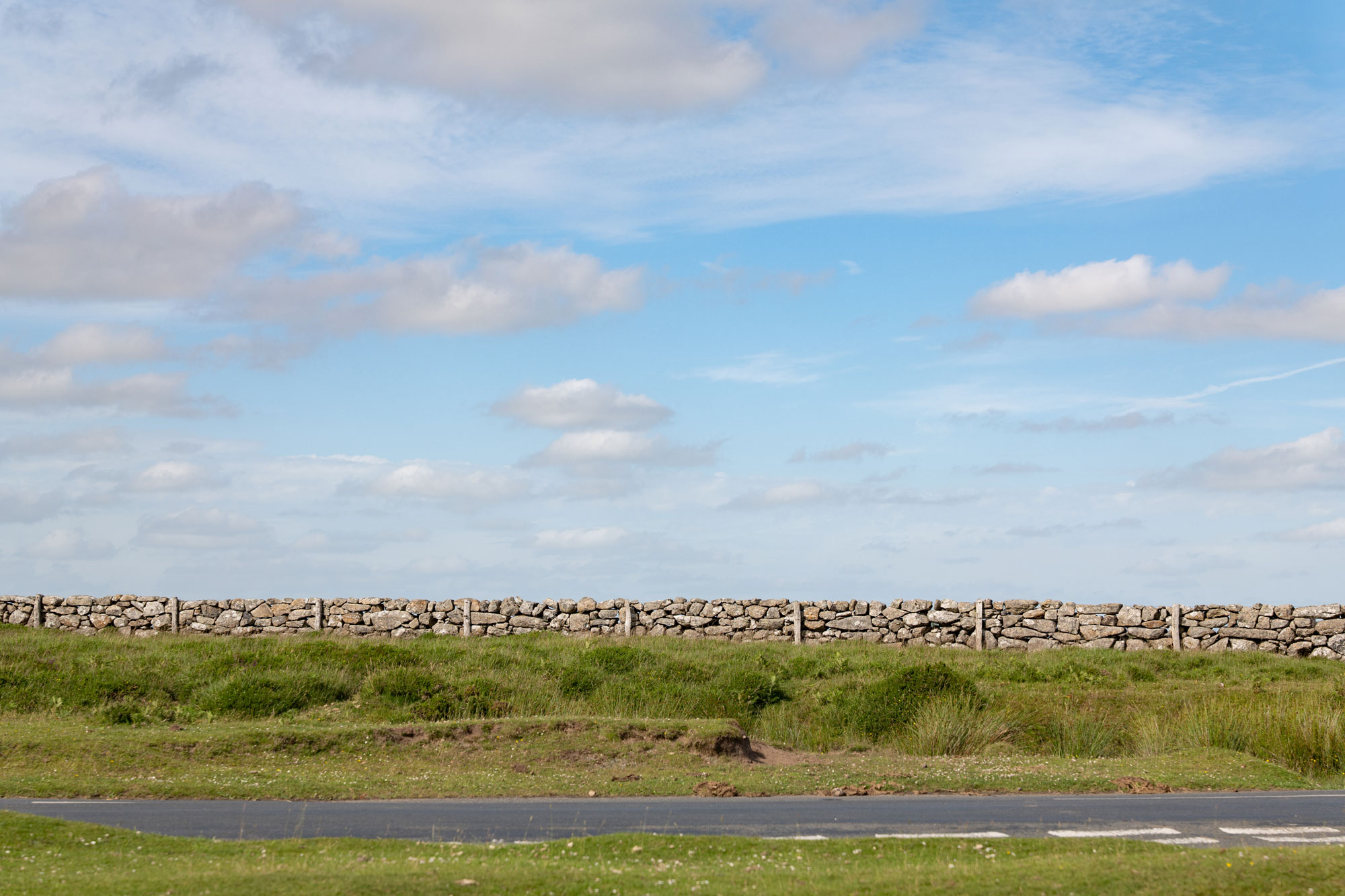A long dry stone wall stretches out across the landscape, with a road in front of it. Blue sky with white clouds can be seen above. Photo by Amy Moore with the Panasonic S1R II.