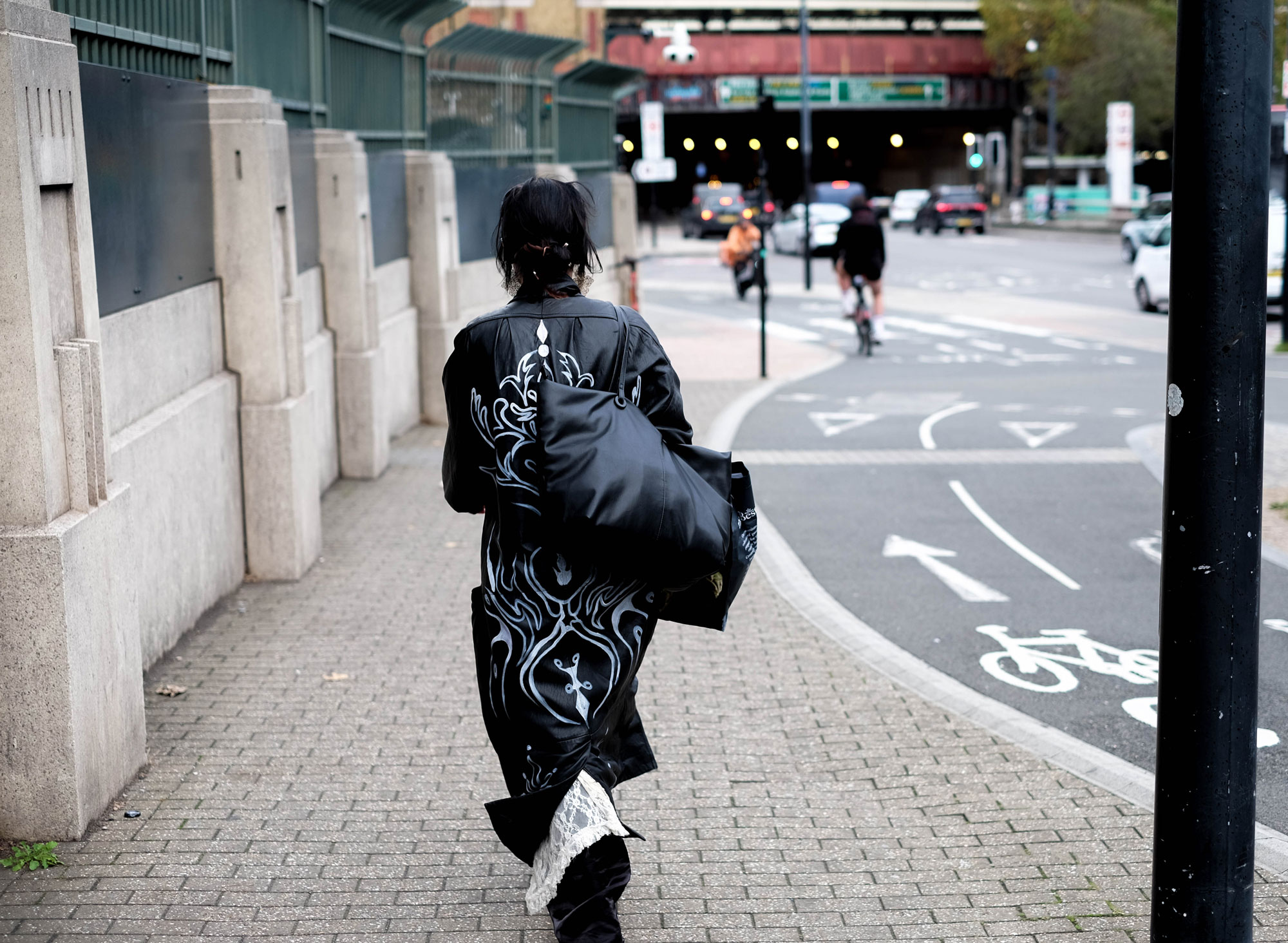 A person wearing a long black trench coat with painted white detailing walks away from the camera. A road with traffic and cyclists is visible in the background.