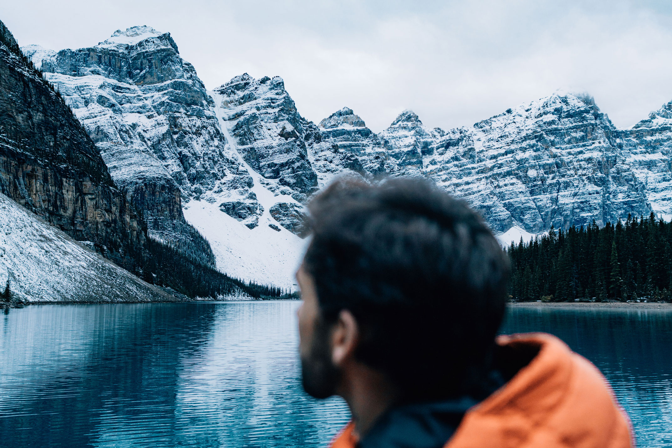 Person in orange jacket in front of Lake Moraine with snow capped mountains in Banff National Park, photo by Jude Allen.