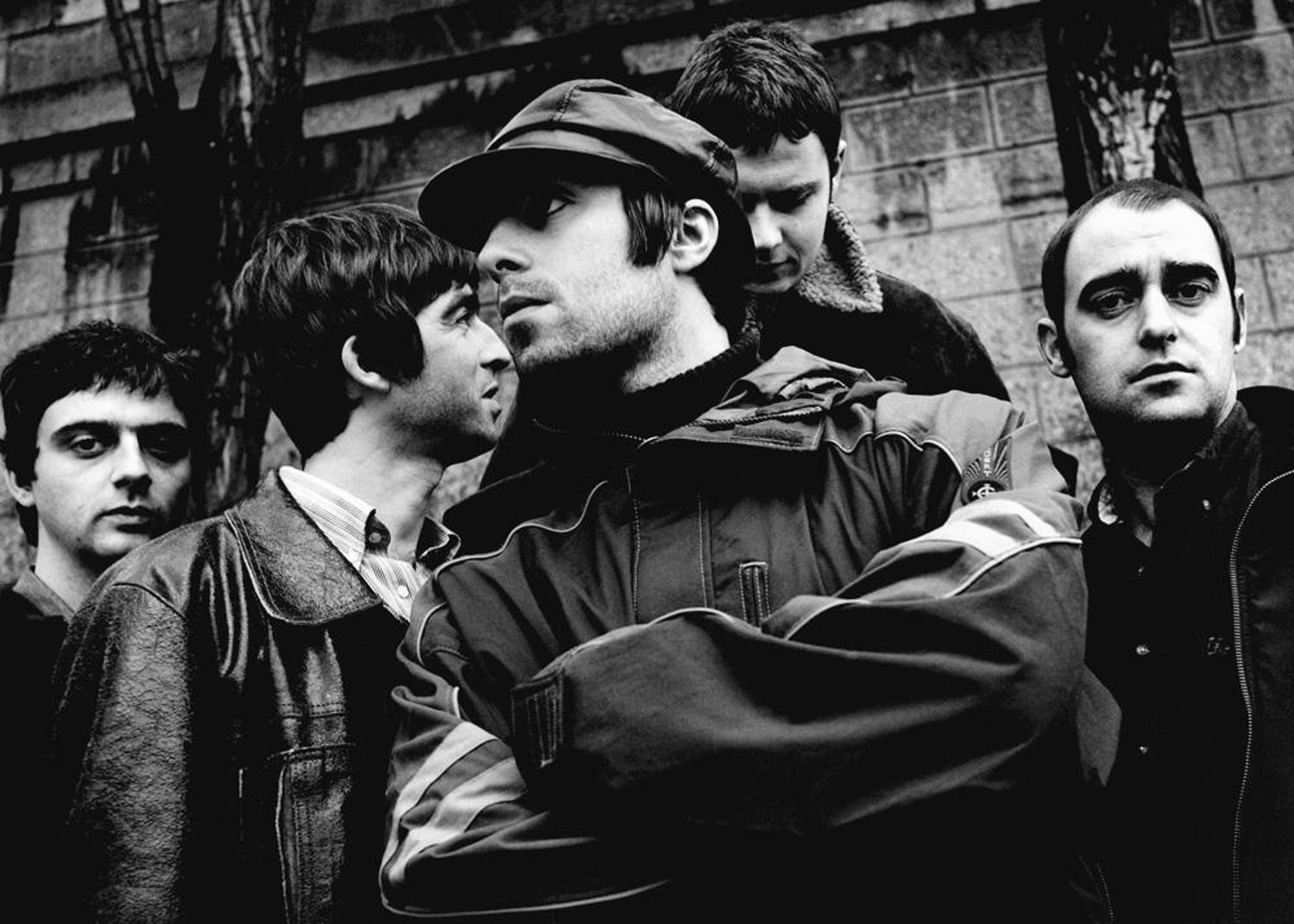 Une photo en noir et blanc de Oasis devant un mur en brique. Photo par Jill Furmanovsky