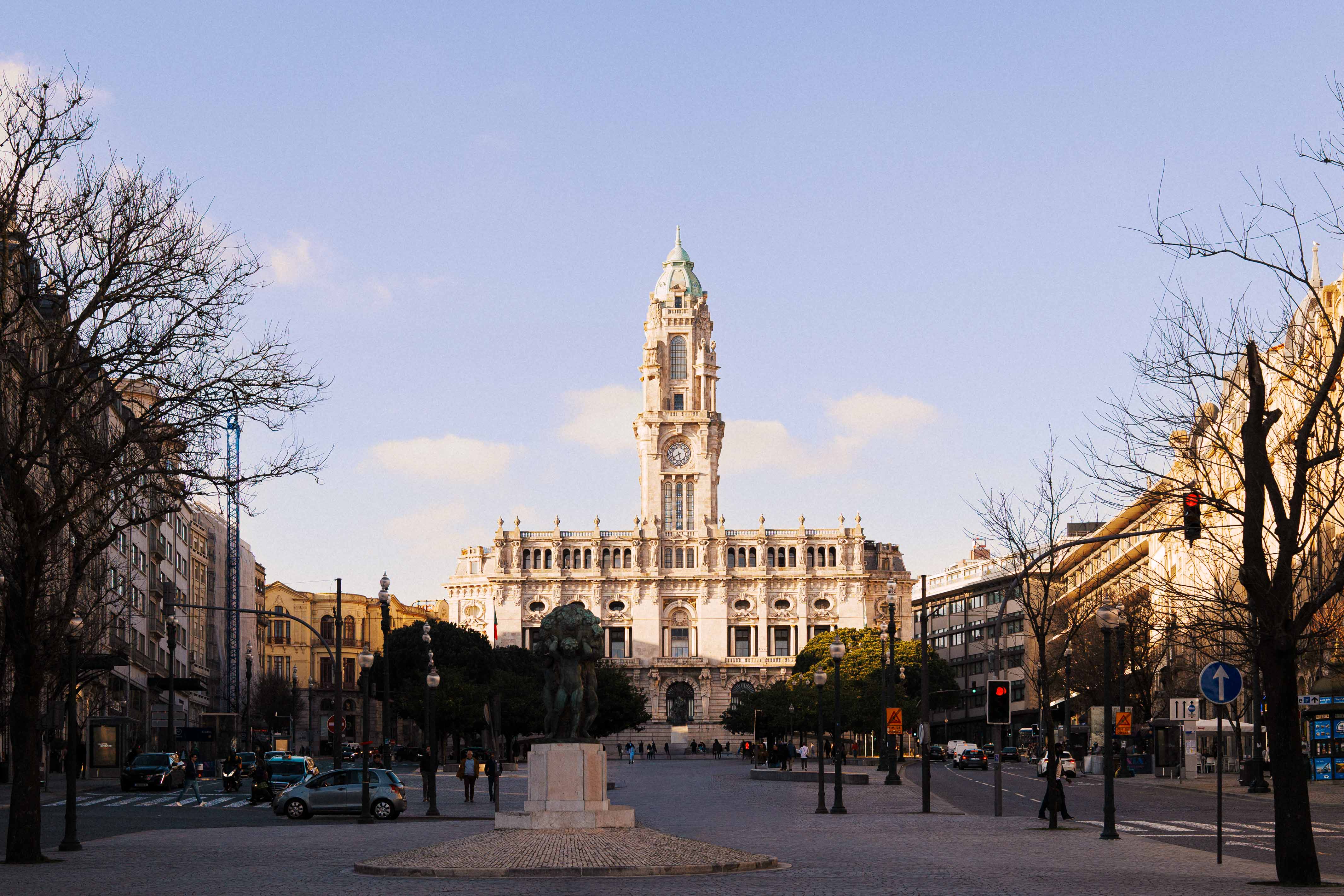 Symmetrical view of Porto City Hall at the end of Avenida dos Aliados, with the clock tower rising above a wide civic square and light traffic passing through in soft afternoon light.

