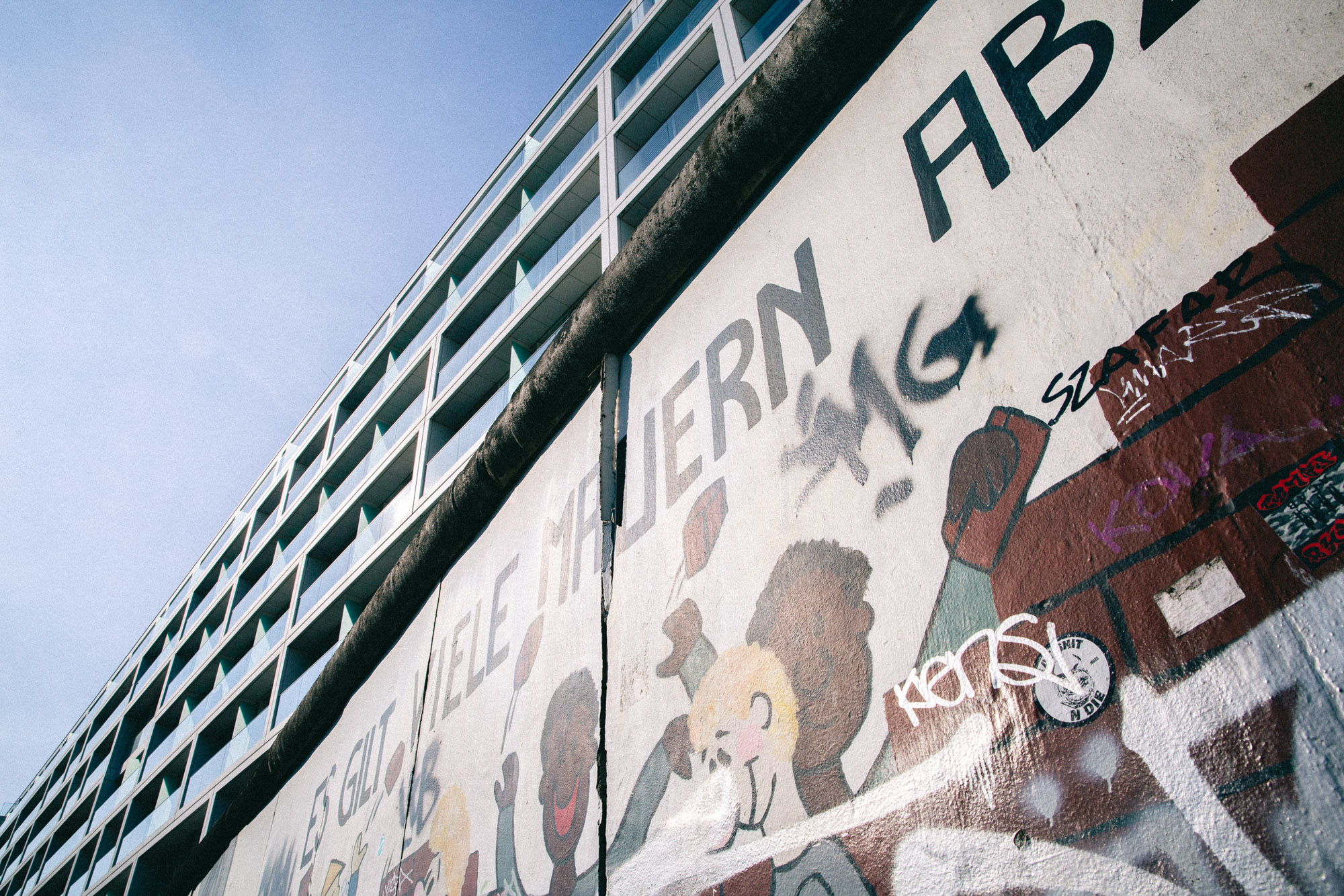 Mural at Berlin’s East Side Gallery with layered graffiti and modern apartment blocks rising in the background. Shot from a low angle in bright daylight by Austin Fassino.