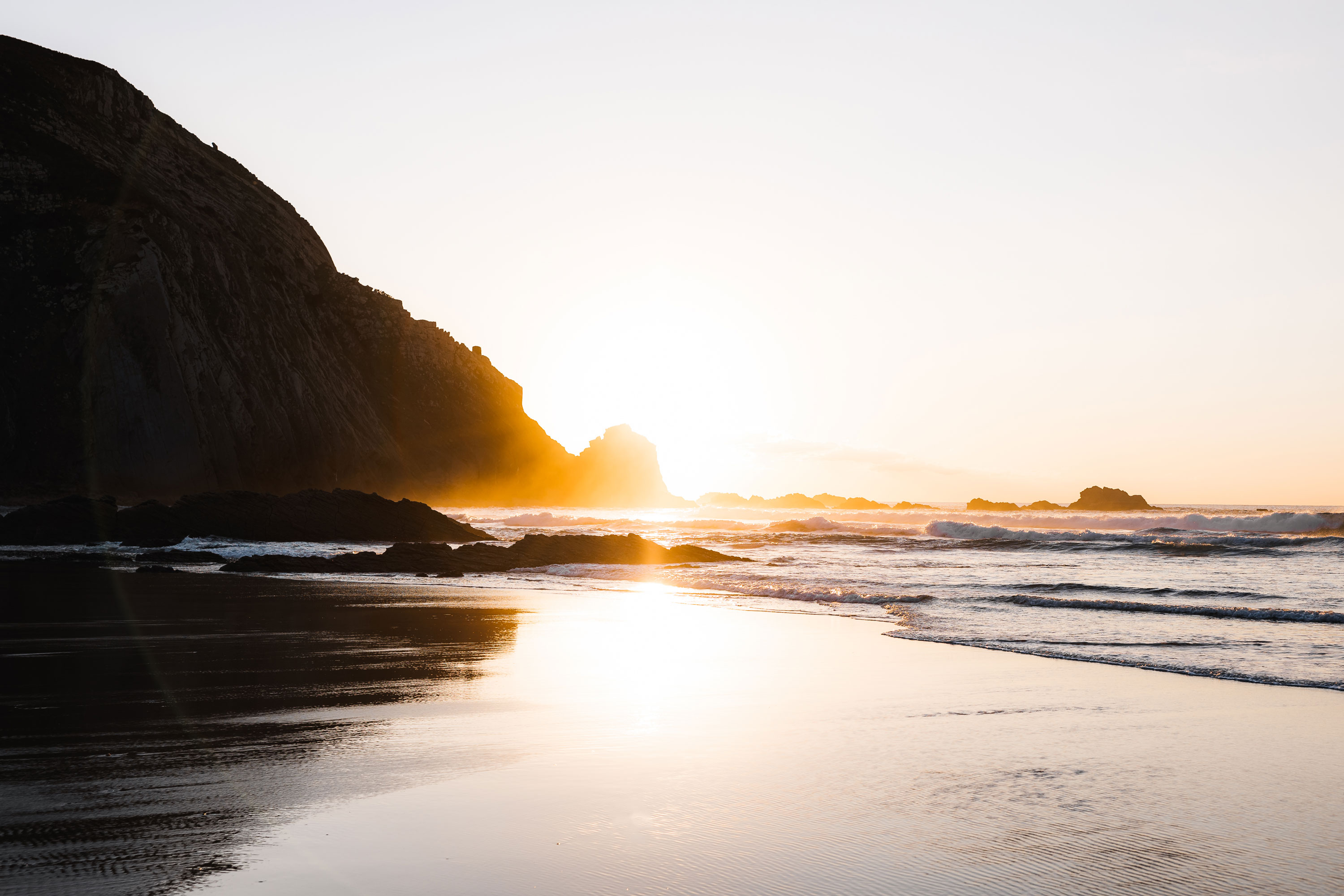 Landscape shot by the sea, mountains in the background with the sun peeking out behind them

