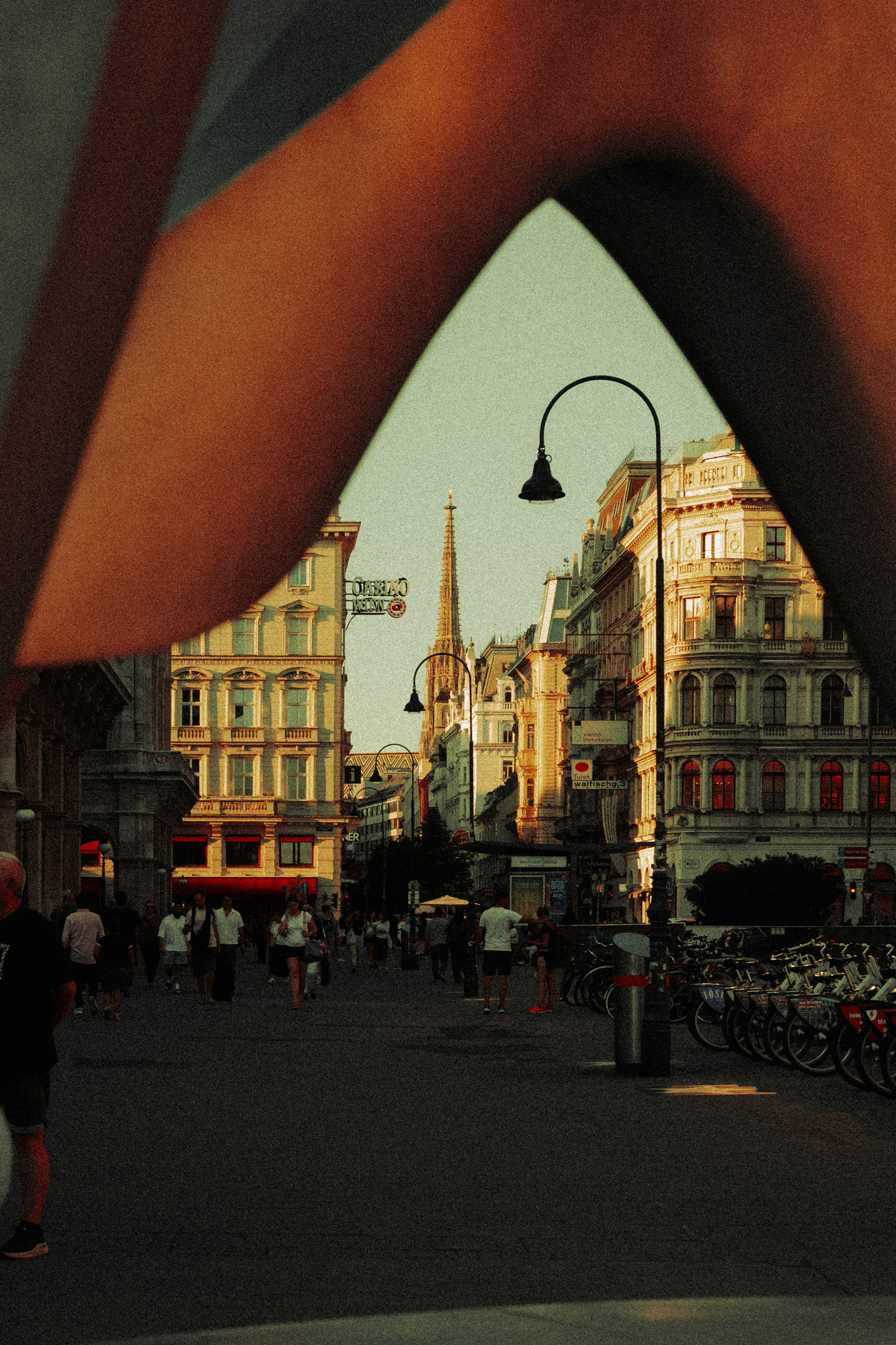 Belebte Straßenszene in Wien. Im Vordergrund rahmen unscharfe Formen den Blick auf Passant:innen, historische Gebäude und einen schlanken Kirchturm in der Ferne. Aufgenommen von Dhan Limwattana mit einer Nikon Z5 II.