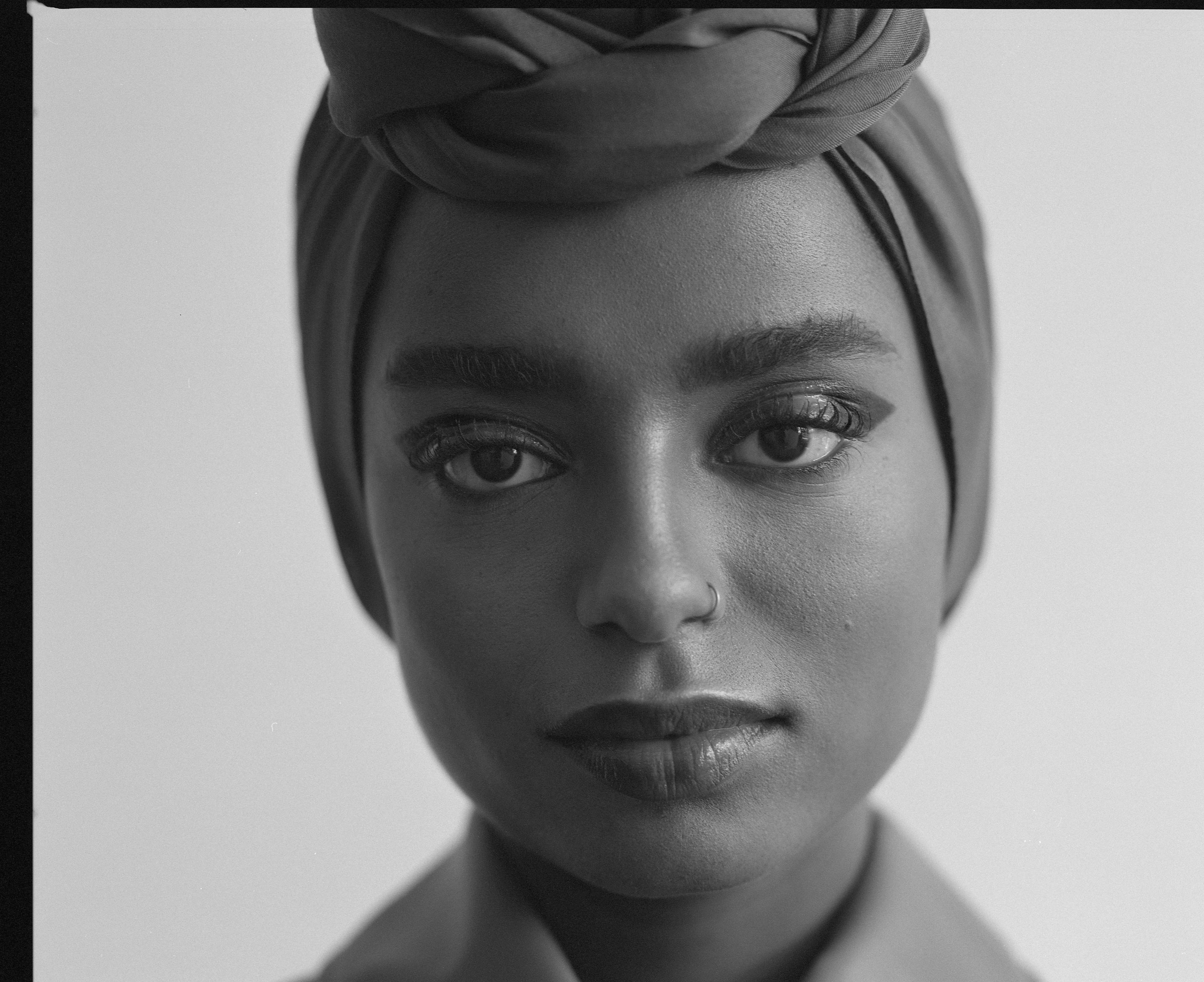 Black and white shot of a Black woman with headscarf and nose ring looking straight at the camera, shot by Samia Rachel.

