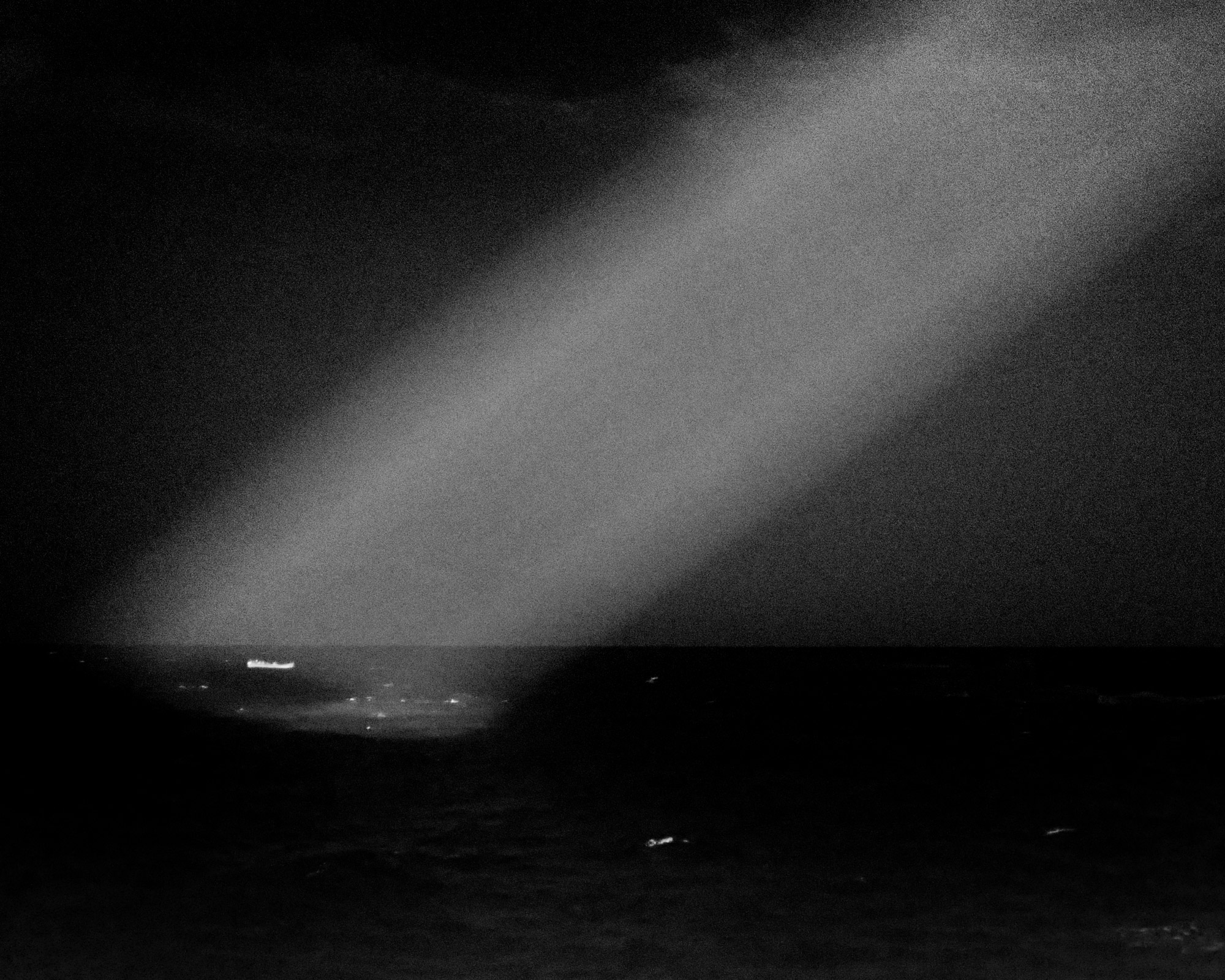 Black and white shot of a small boat in the sea, directly illuminated from the top right.