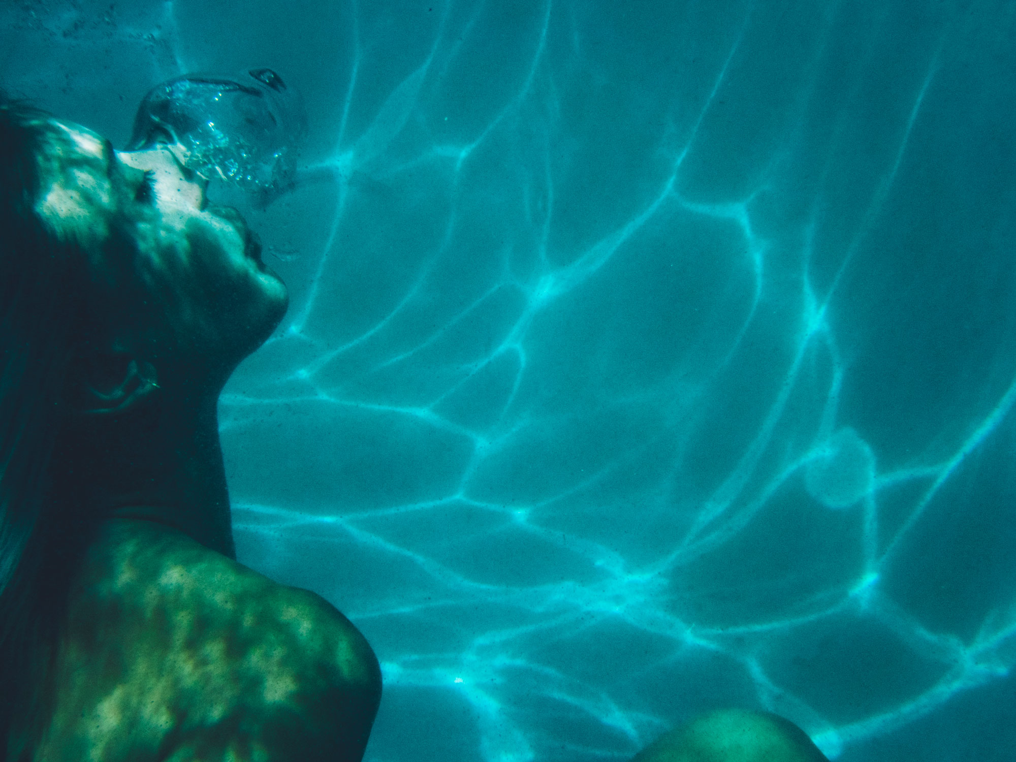 Underwater image of a woman in a pool blowing a bubble by Lindsey Byrnes.