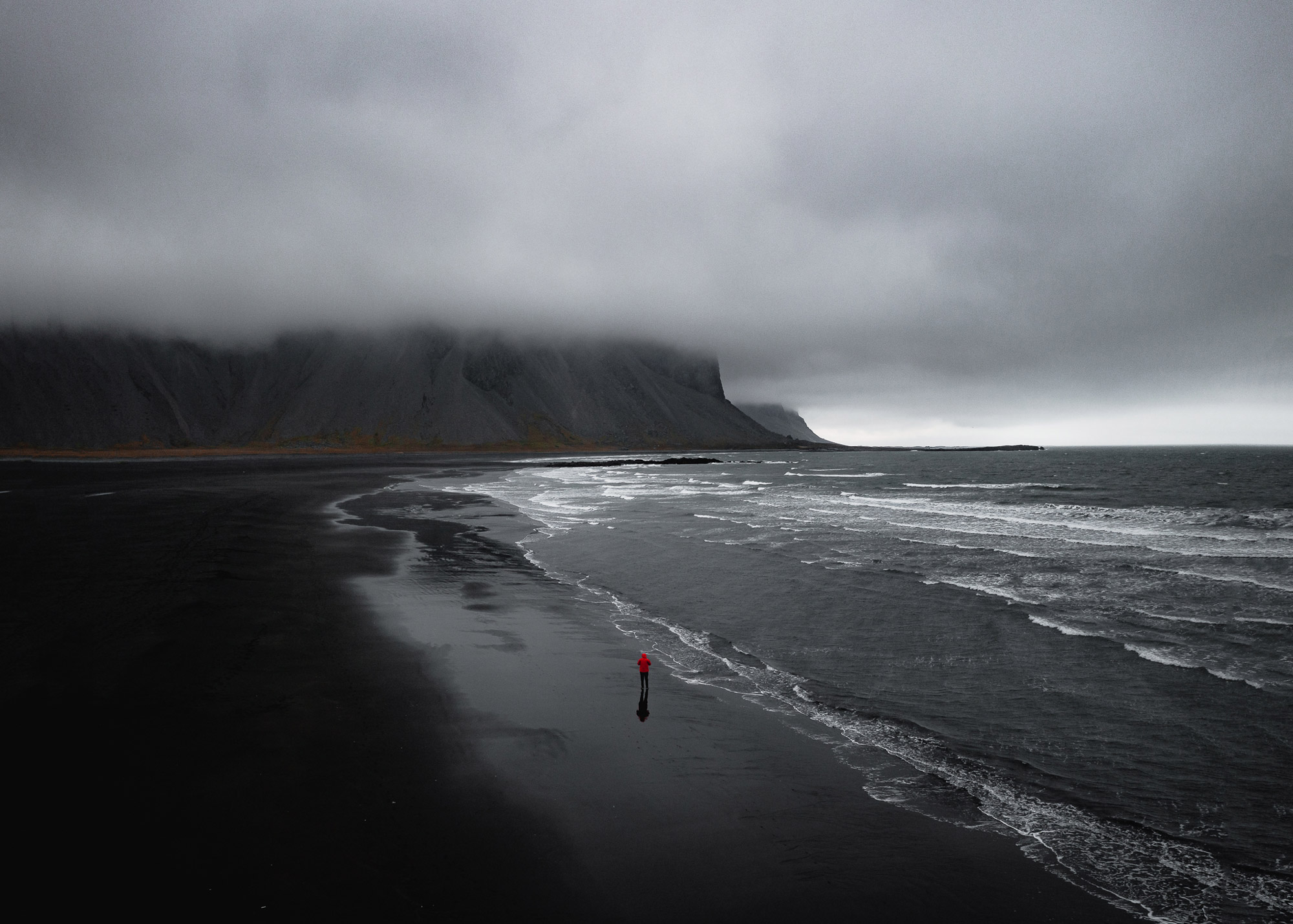 An aerial drone photo of a figure in a red coat walking down a dark moody coastline in Iceland, there are hills and clouds in the background, shot by Paul Watson with a DJI Mavic 2 Pro