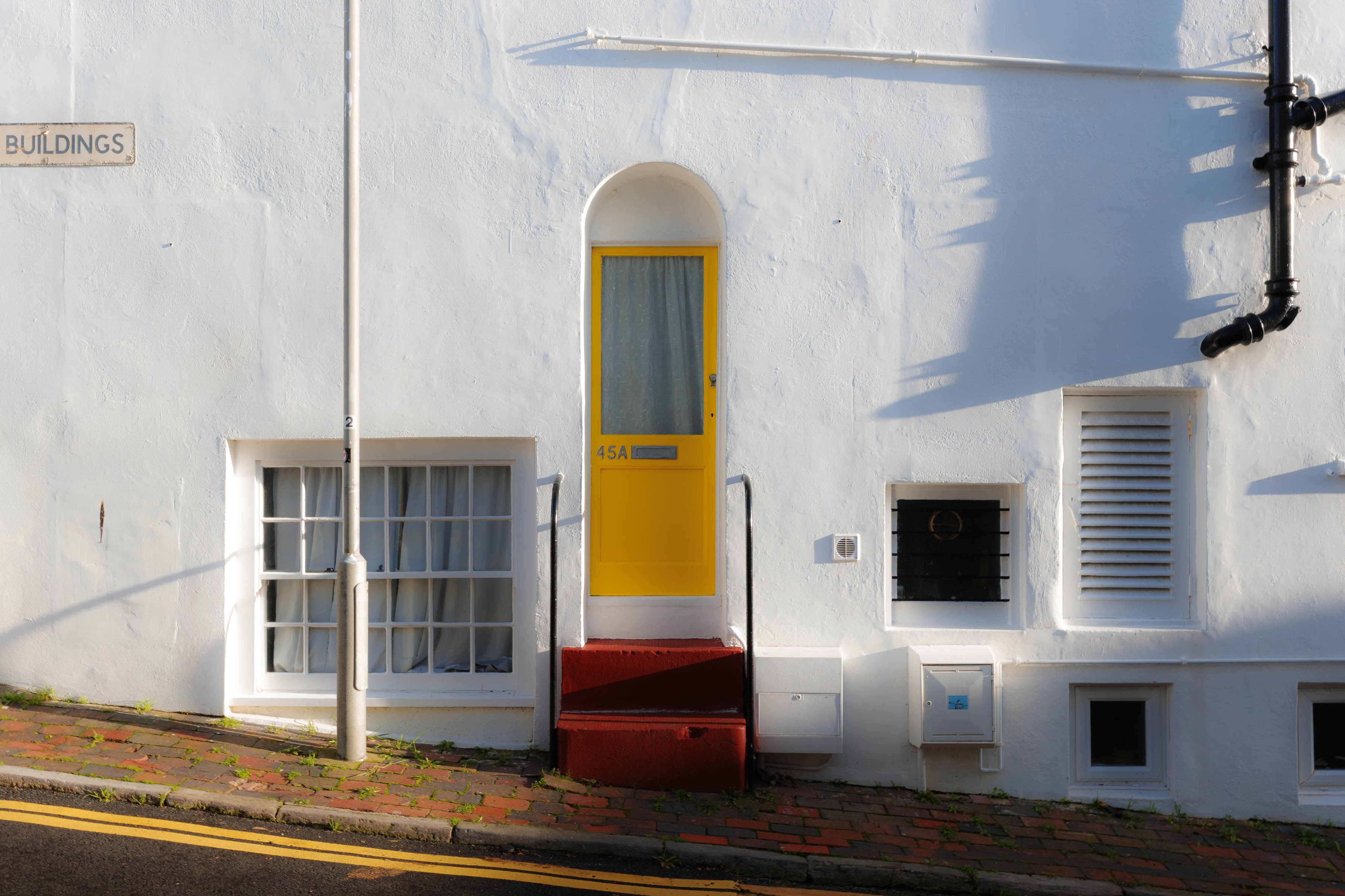 Ein sauberes, minimalistisches Foto eines weiß gestrichenen Gebäudes mit einer leuchtend gelben Tür, roten Stufen und architektonischen Details bei weichem Tageslicht. Aufgenommen mit einer Ricoh GR IV.