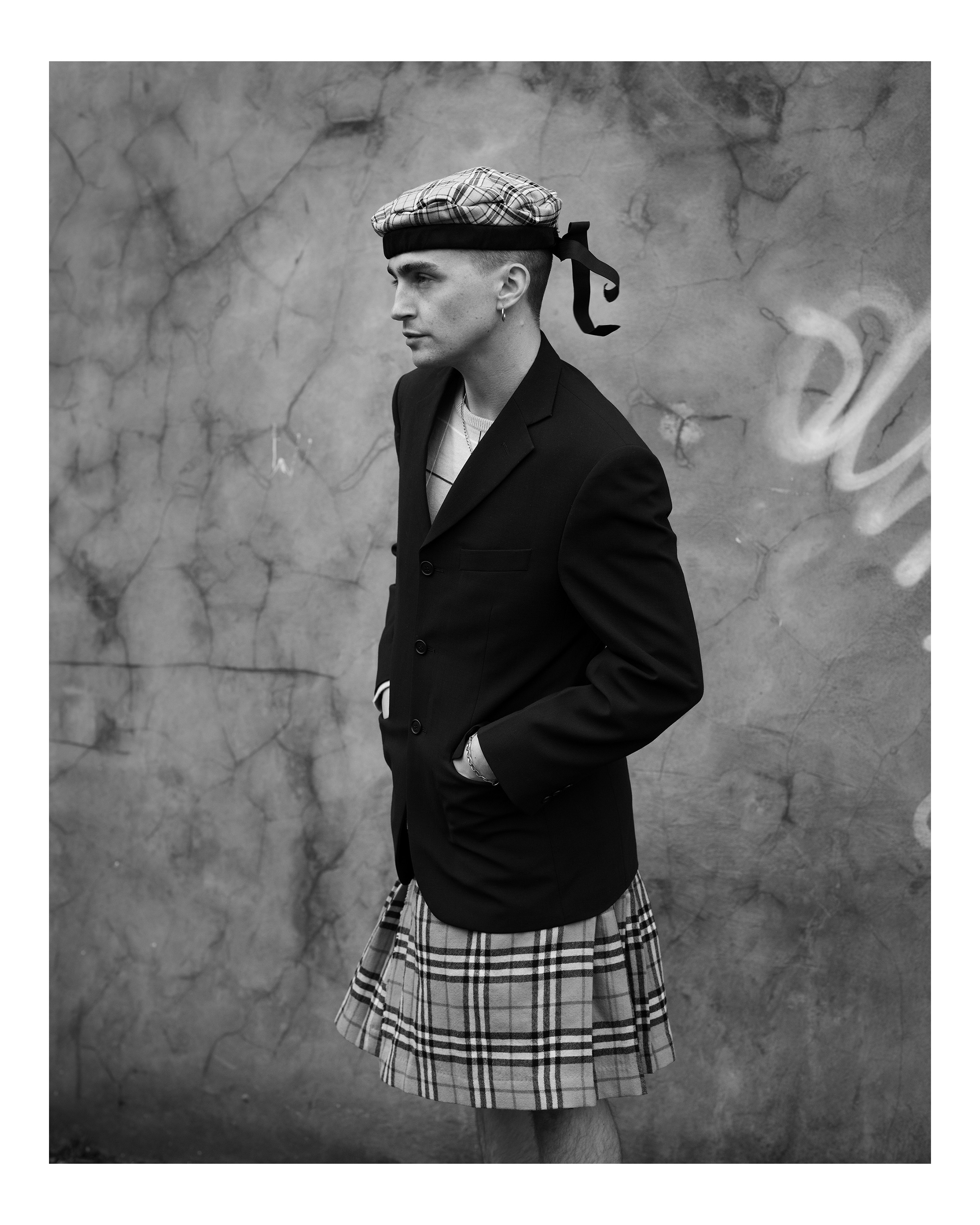 Schwarz-weißes Oberkörperporträt eines Mannes, der einen Kilt, einen Tartan-Tam o' Shanter-Hut und einen Blazer trägt und vor einer grauen Wand steht, fotografiert von Simon Murphy mit einer Fujifilm GFX 100.