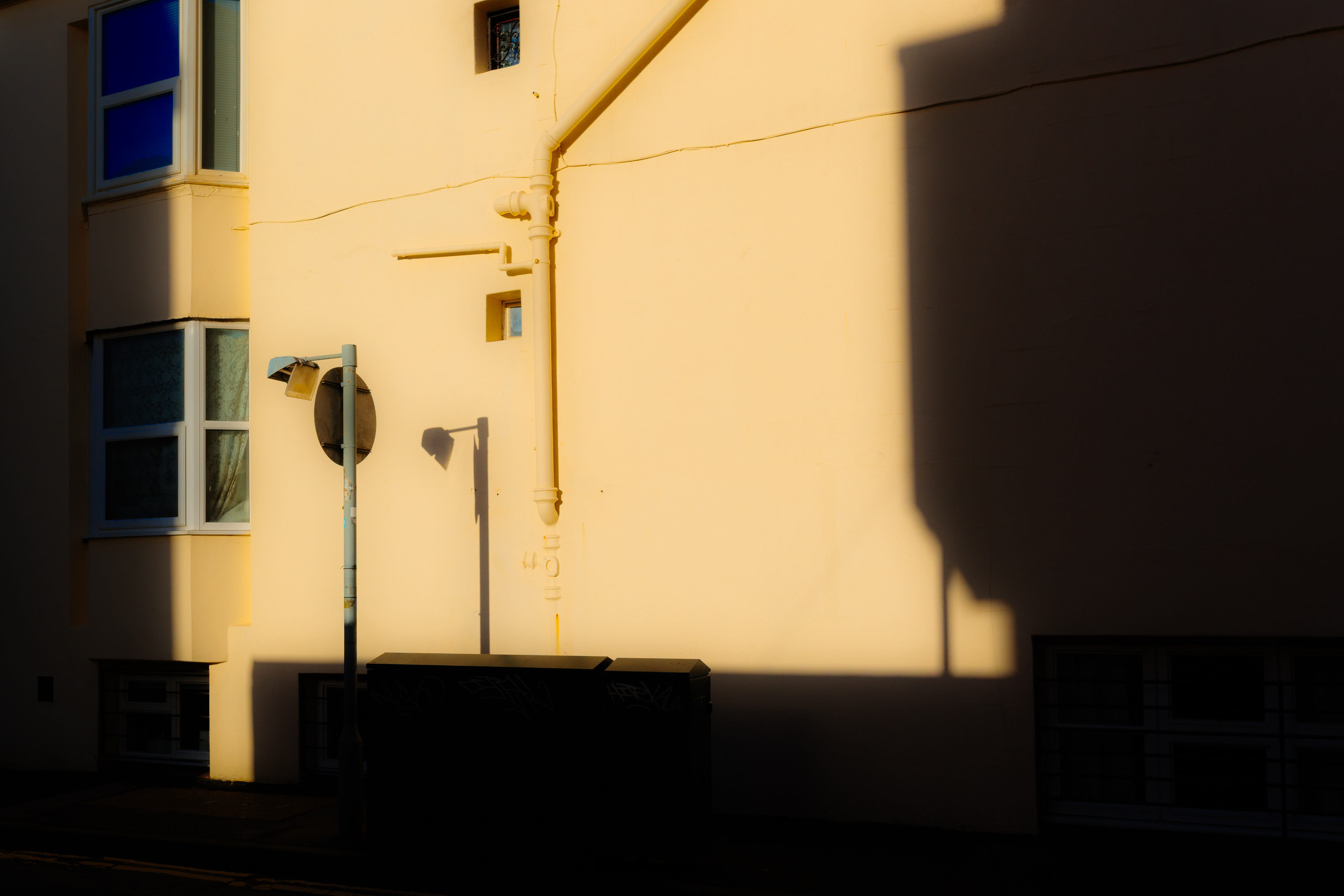 A minimalist street photograph showing a sunlit cream-coloured building facade with deep shadows, windows and street signage, captured in warm golden-hour light with the Ricoh GR IV compact camera.