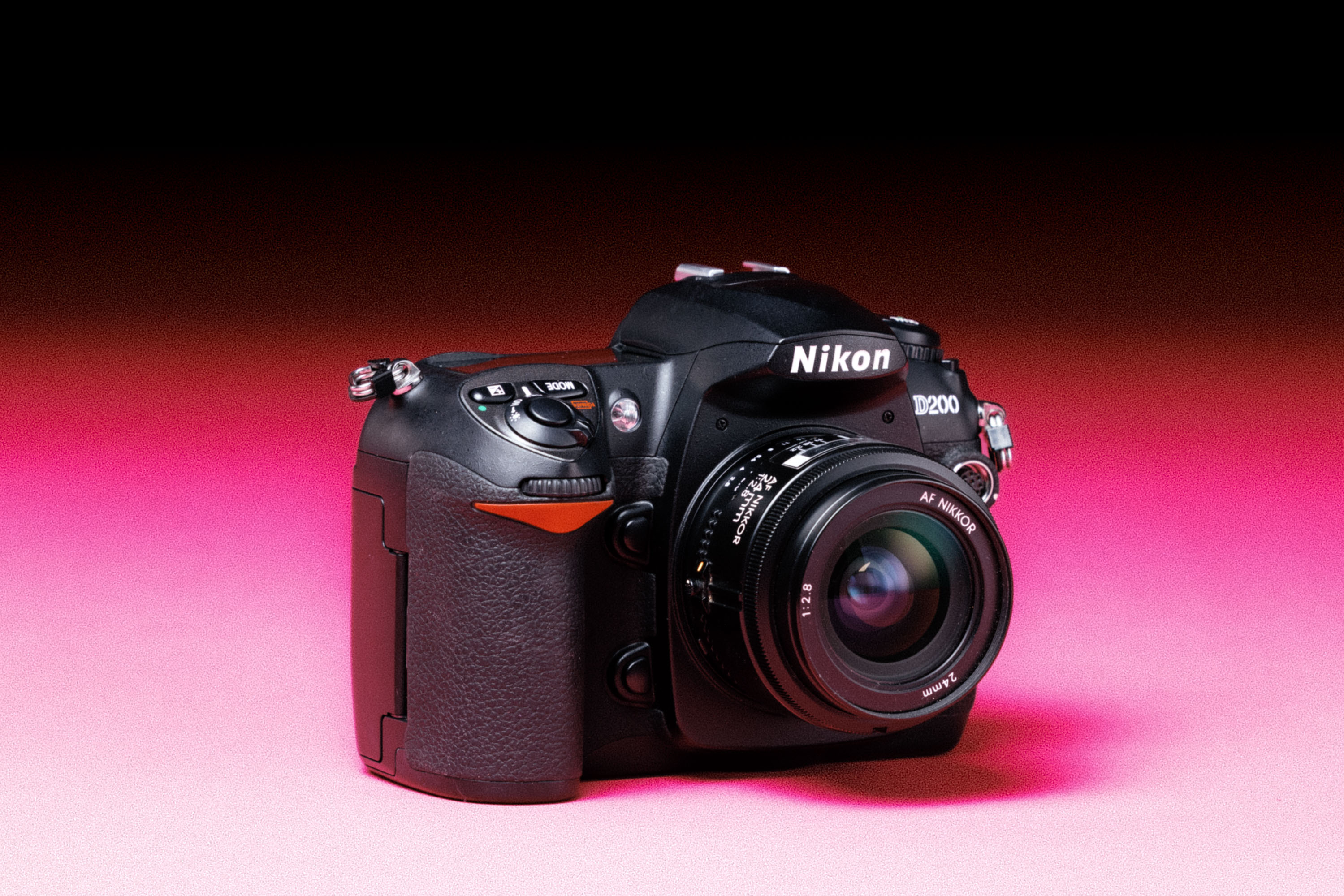 A Nikon D200 camera with a Nikon AF Nikkon 24mm f/2.8D lens, with an added film-like grain effect on a graduated pink background. 
