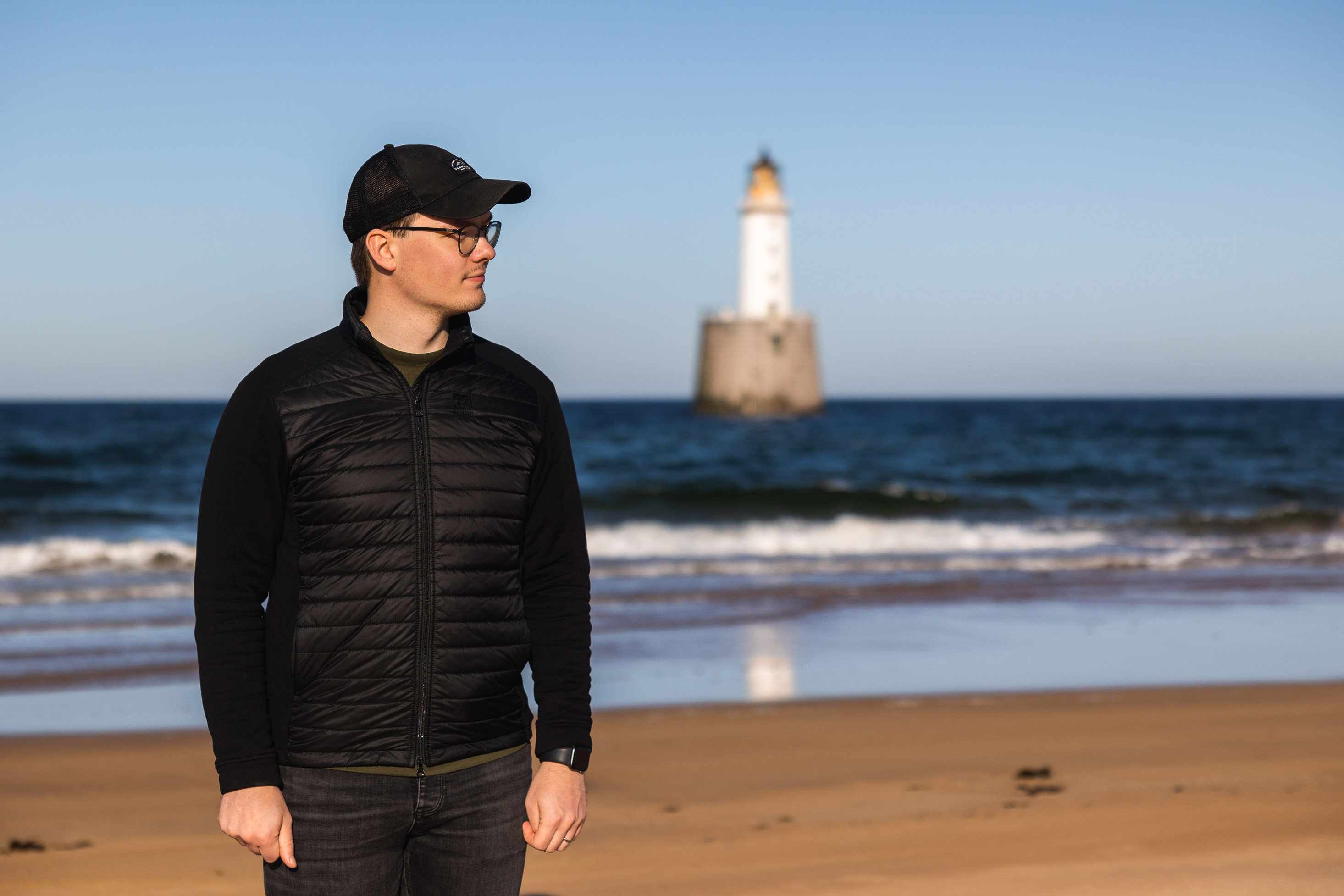 a man wearing a hat standing on a beach with the sea and a lighthouse in the background