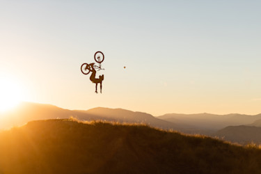 Silueta de un ciclista haciendo un salto mortal al atardecer sobre una cadena montañosa.