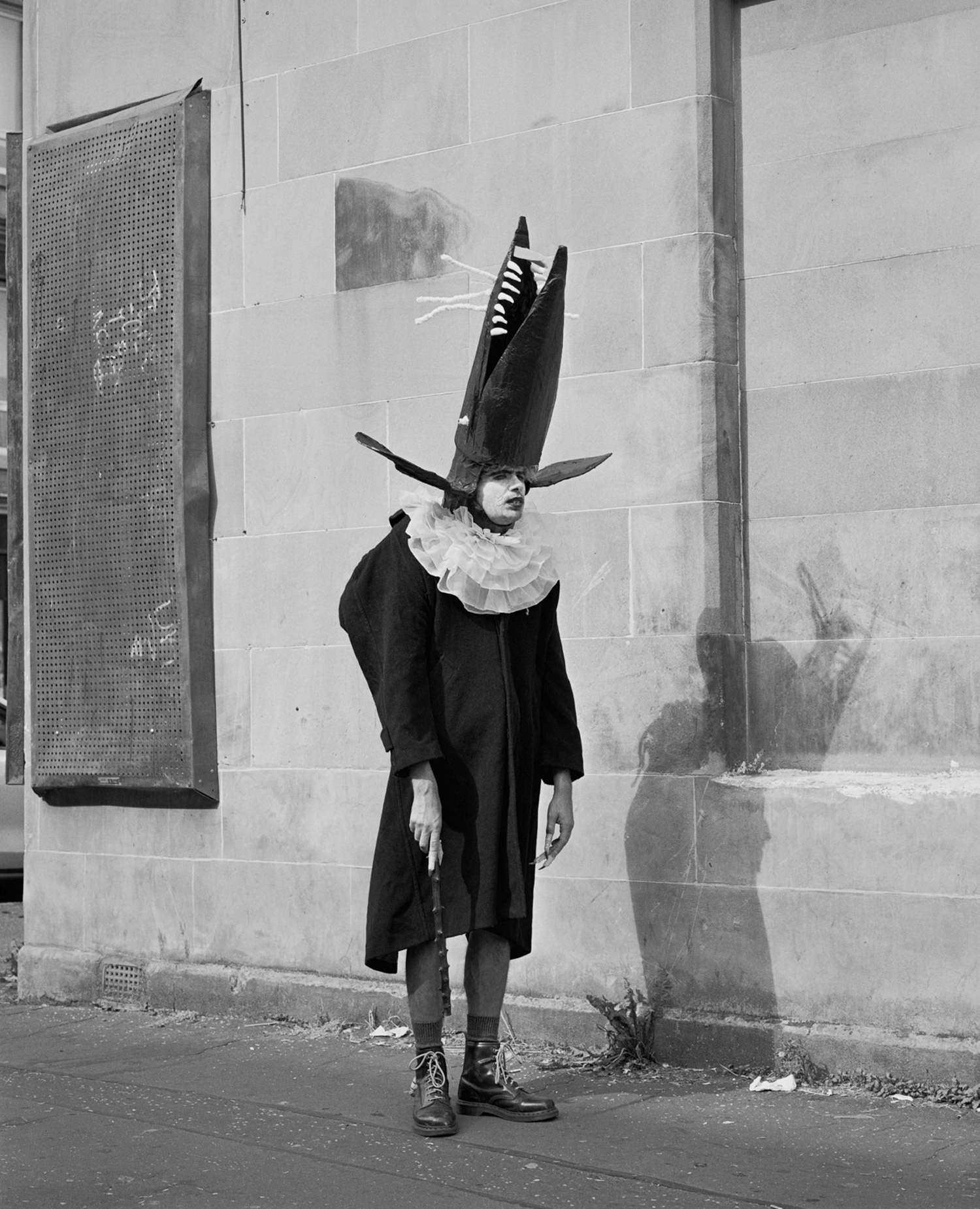 Black-and-white street portrait of a person in theatrical costume of a rat outside an old building, photo by Simon Murphy
