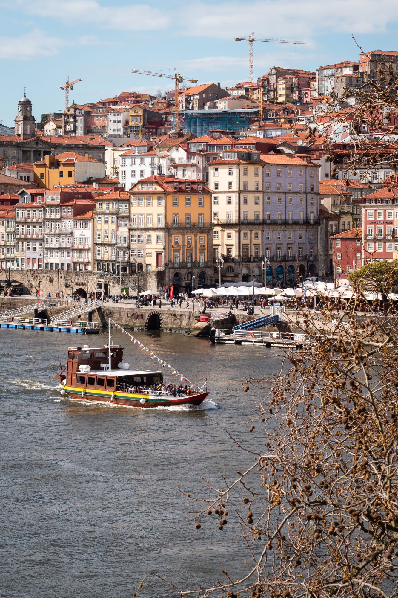 Ein fahrendes Boot mit verschiedenen Länderflaggen. Im Hintergrund eine belebte Uferpromenade mit weißen Sonnenschirmen und mehreren Kränen.