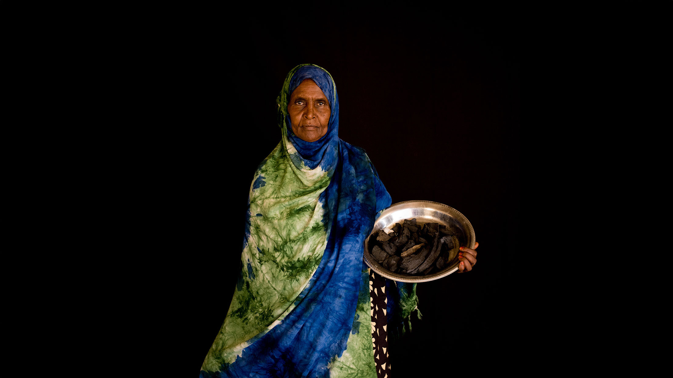 A brightly dressed woman in green and blue carrying a silver bowl with food in it.