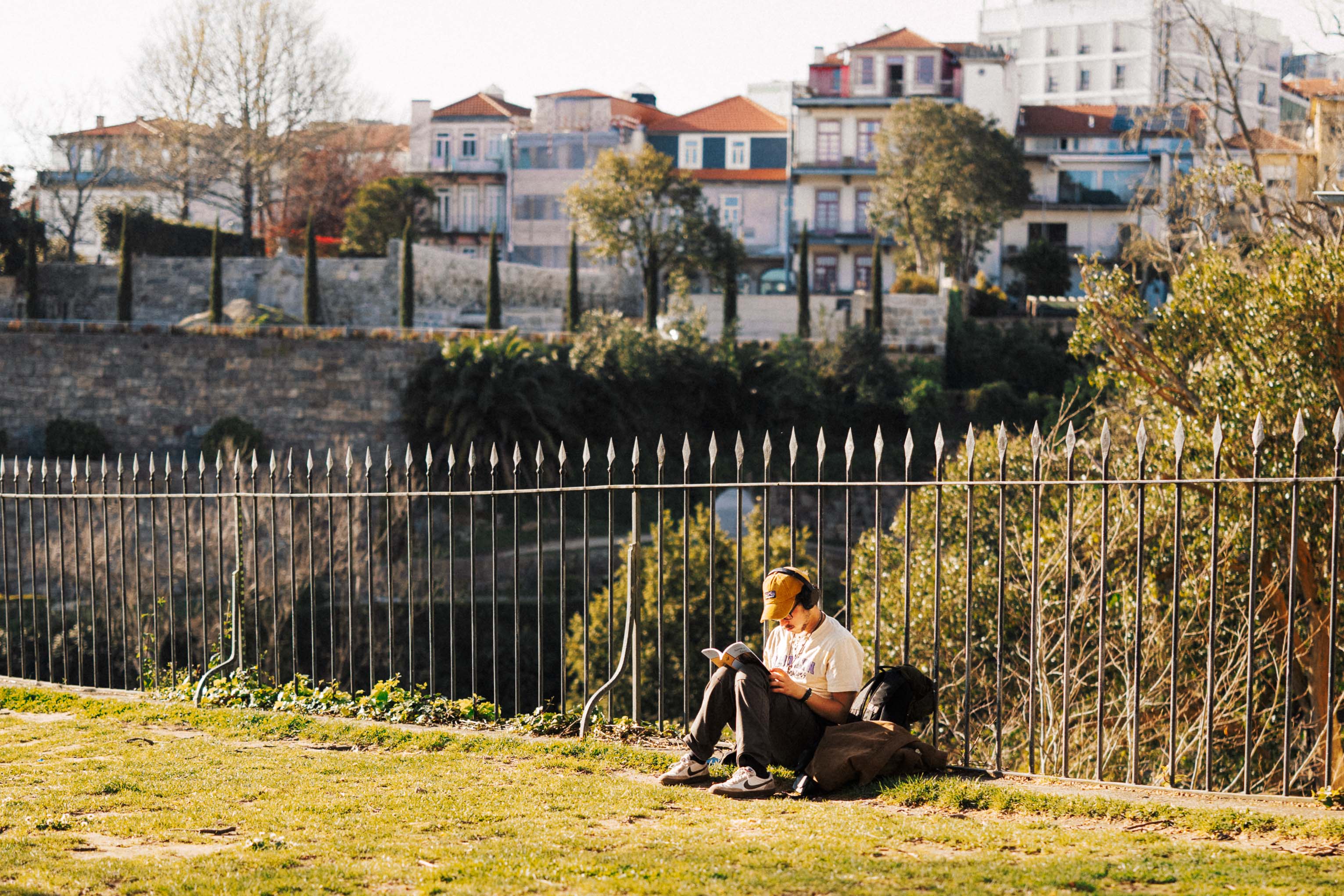 A person sitting on the grass reading a book at Passeio das Virtudes Gardens, with a metal fence in the foreground and hillside houses and trees overlooking the Douro Valley in soft afternoon light.
