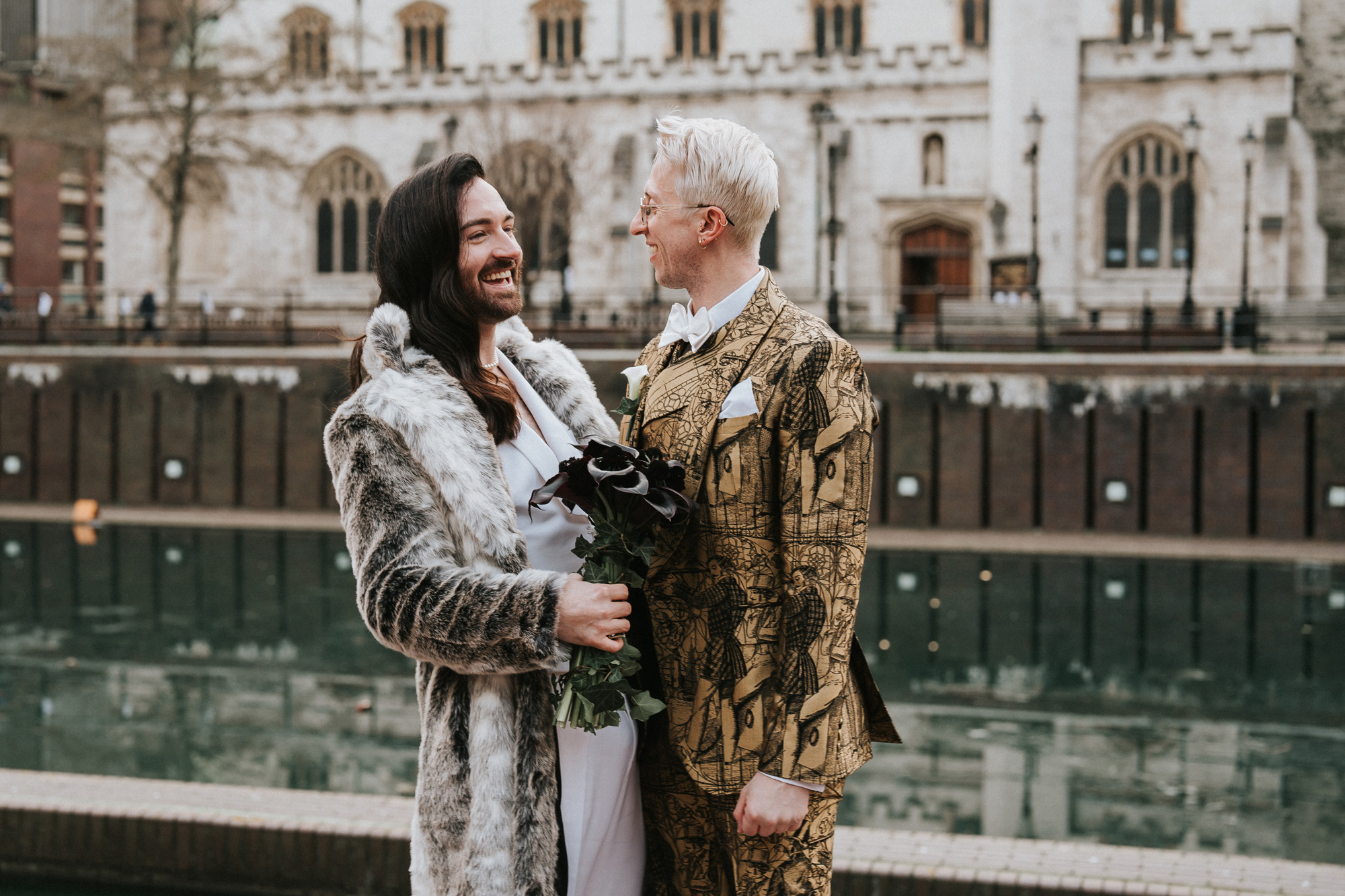 Des jeunes mariés célèbrent leur mariage près d'un canal, à l'extérieur d'une cathédrale. L'un porte un manteau de fourrure et tient un bouquet de fleurs noires, tandis que l'autre porte un costume doré. Tous deux se sourient. Photo prise par Steph Large.