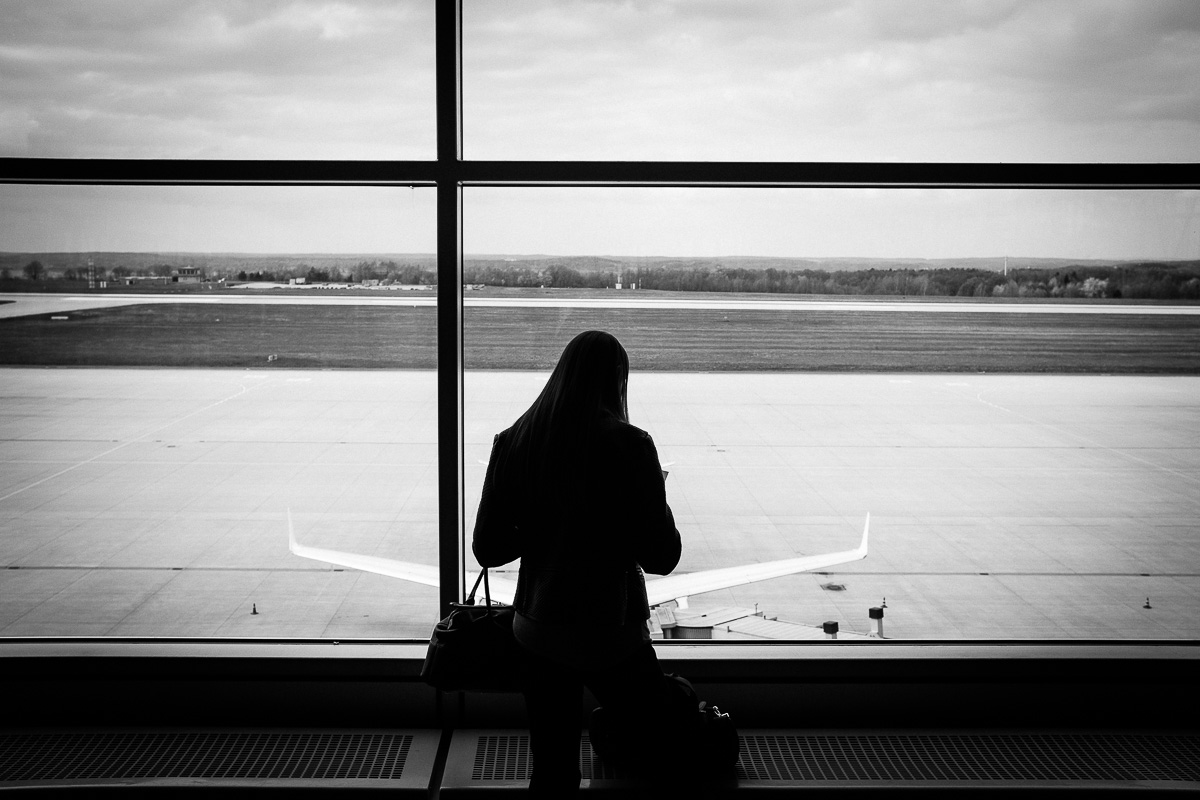 A silhouette of a person, looking down in front of an airport runway, with the wings of a plane in the background appearing from either side of their silhouette, black-and-white street photography by Marco Larousse