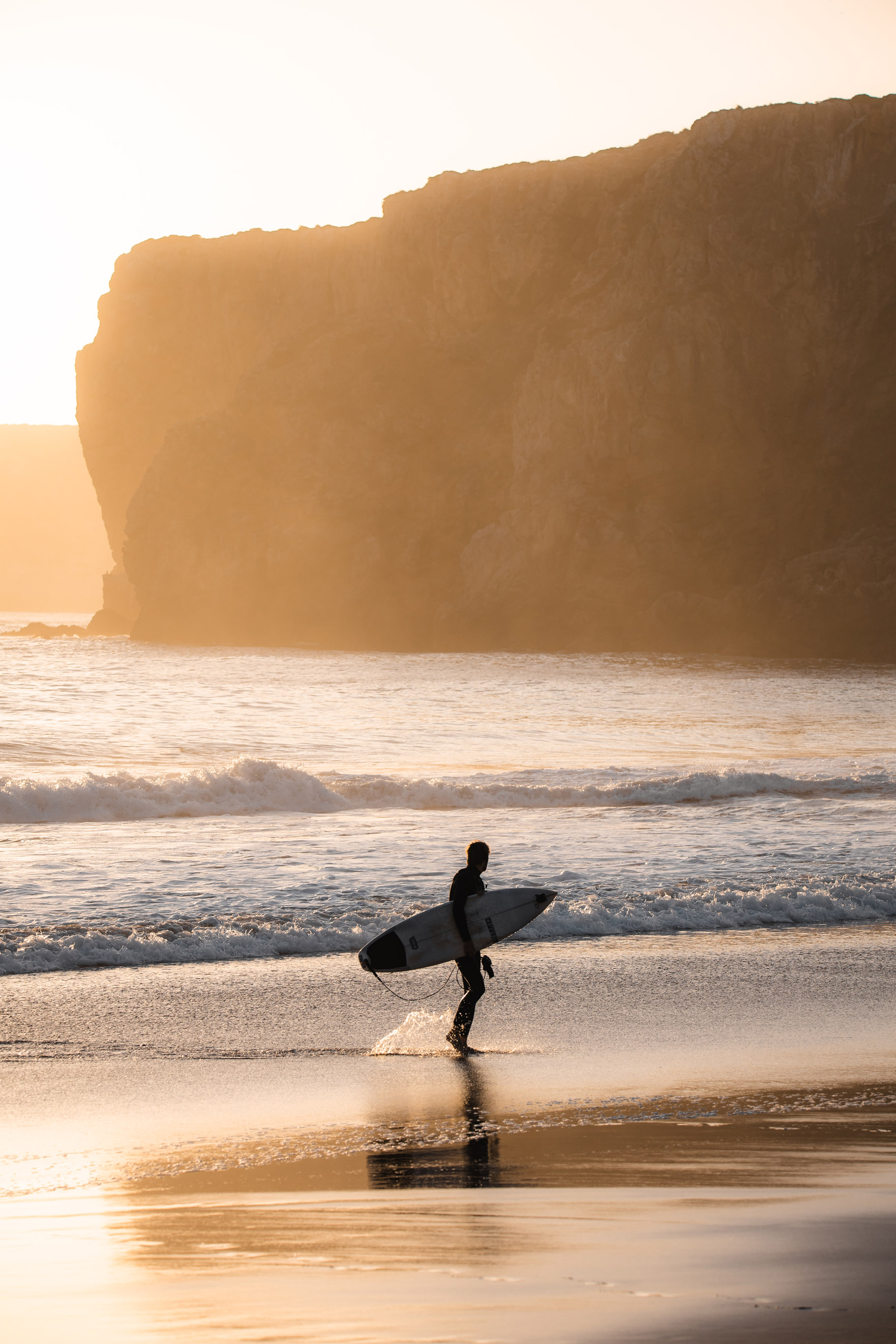 Bild eines Surfers am Strand vor einer Bergkulisse im Abendlicht.