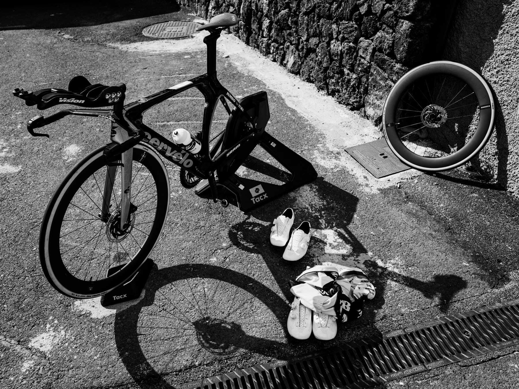 Vélo de course entouré de chaussures de cyclisme et d’une roue de rechange dans une scène en noir et blanc qui met en lumière les préparatifs et les coulisses de la compétition. Photo : Dan King, réalisée avec un Fujifilm X-Pro 3.