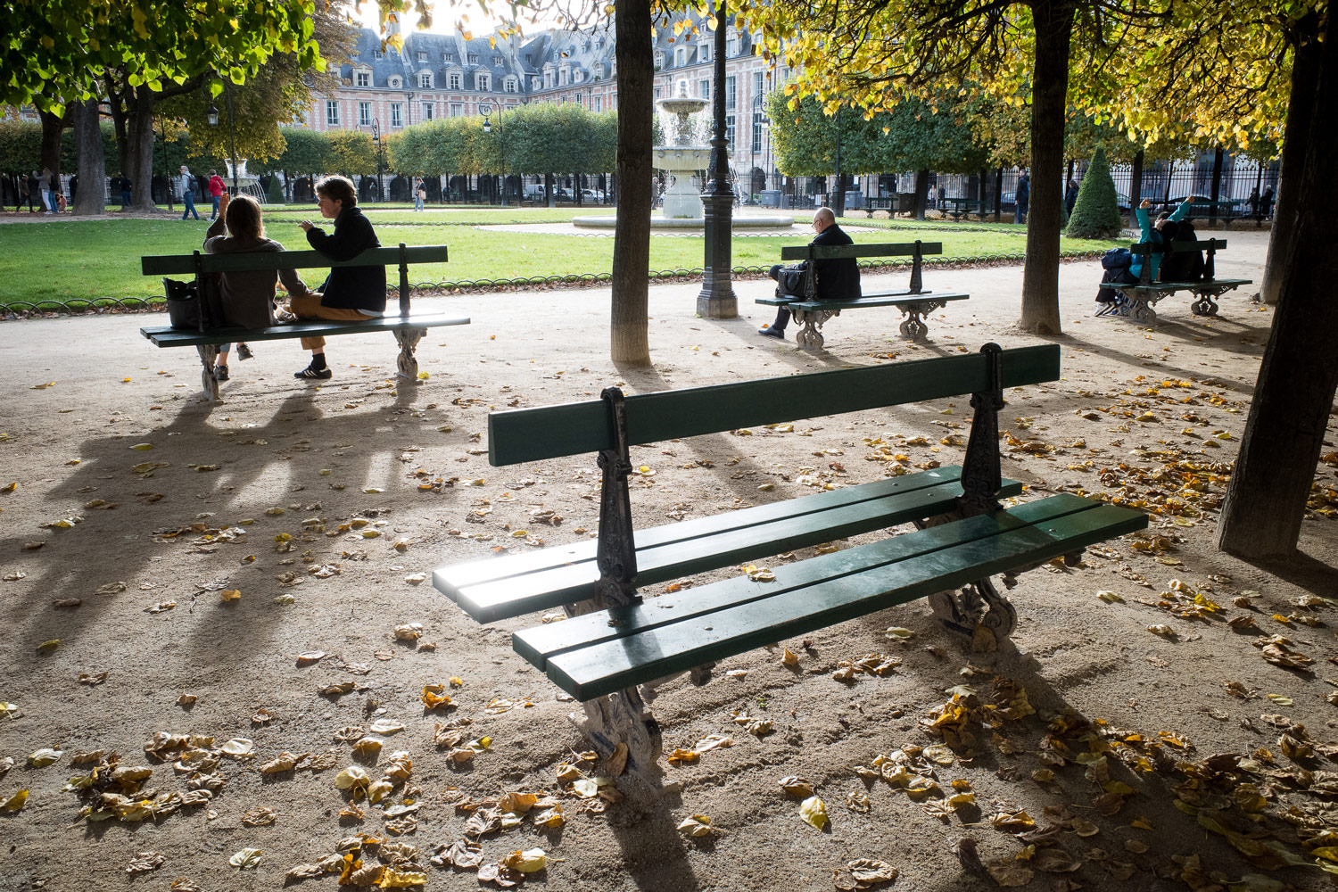 Sunlit benches in Place des Vosges, Paris, surrounded by autumn leaves and trees, with people relaxing near a central fountain in a peaceful city park.
