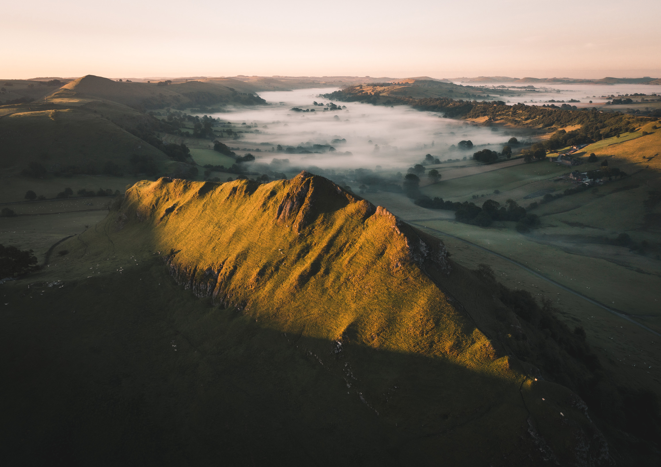 Eine Drohnenaufnahme von Parkhouse Hill, England, mit Nebel in der Ferne, die Sonne beleuchtet die Spitze des nächsten Hügels mit weiteren Hügeln in der Ferne. Aufgenommen von Jordan Lancaster-Graham mit einer DJI Mavic II Pro.