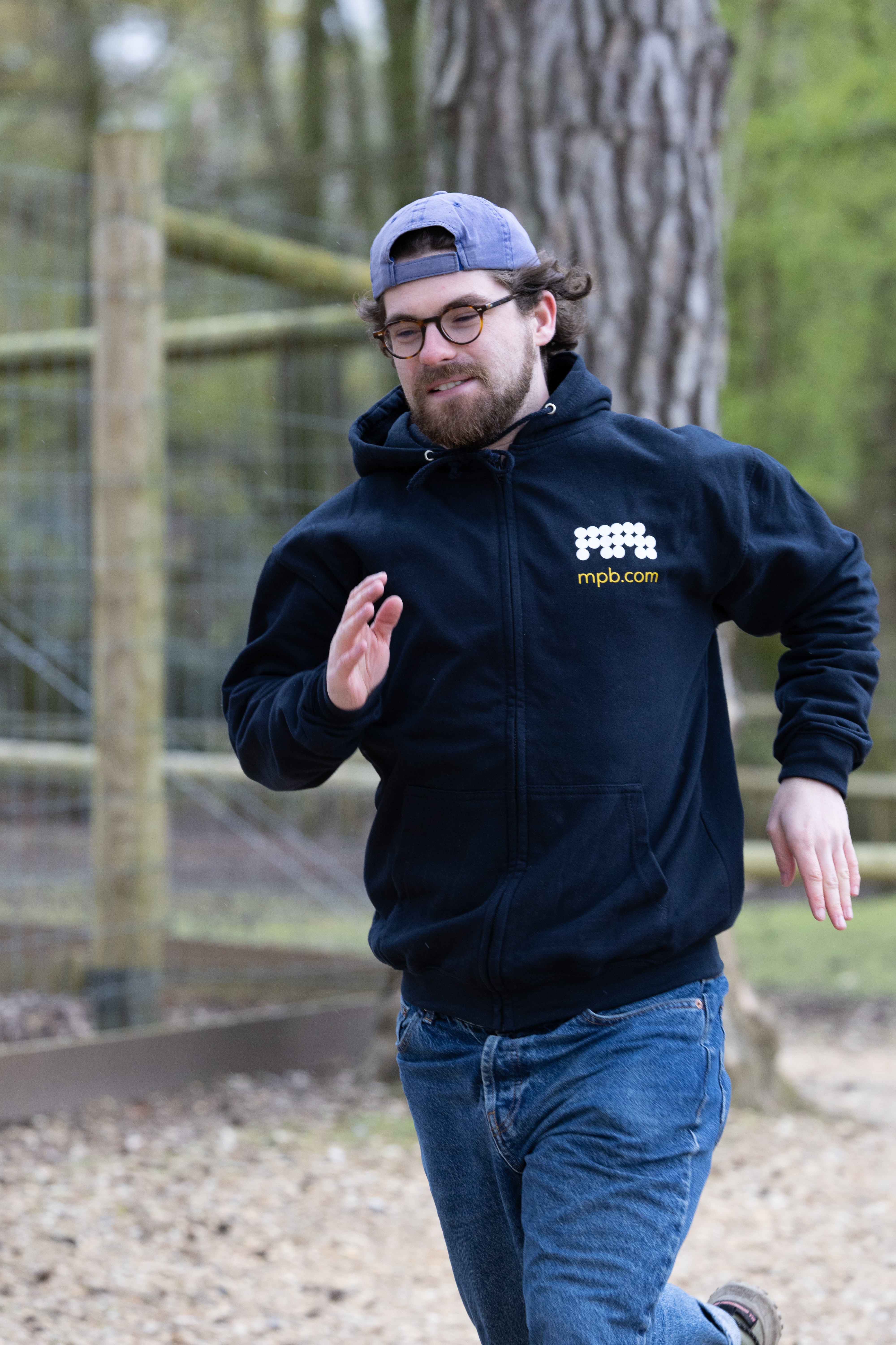 A person mid-sprint, running to the left of the camera. The person is wearing a cap and glasses, as well as a zip-up hoodie with the old MPB logo.