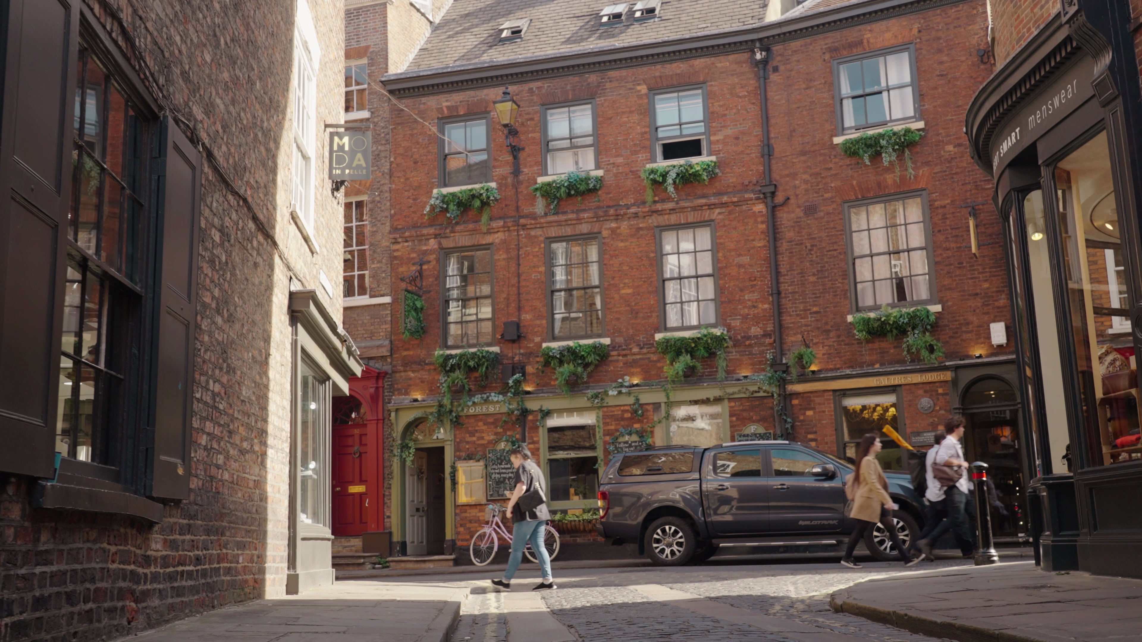 A traditional red-brick pub in York with foliage growing from the windowsills, a cyclist is walking past, a car is parked in front of the pub