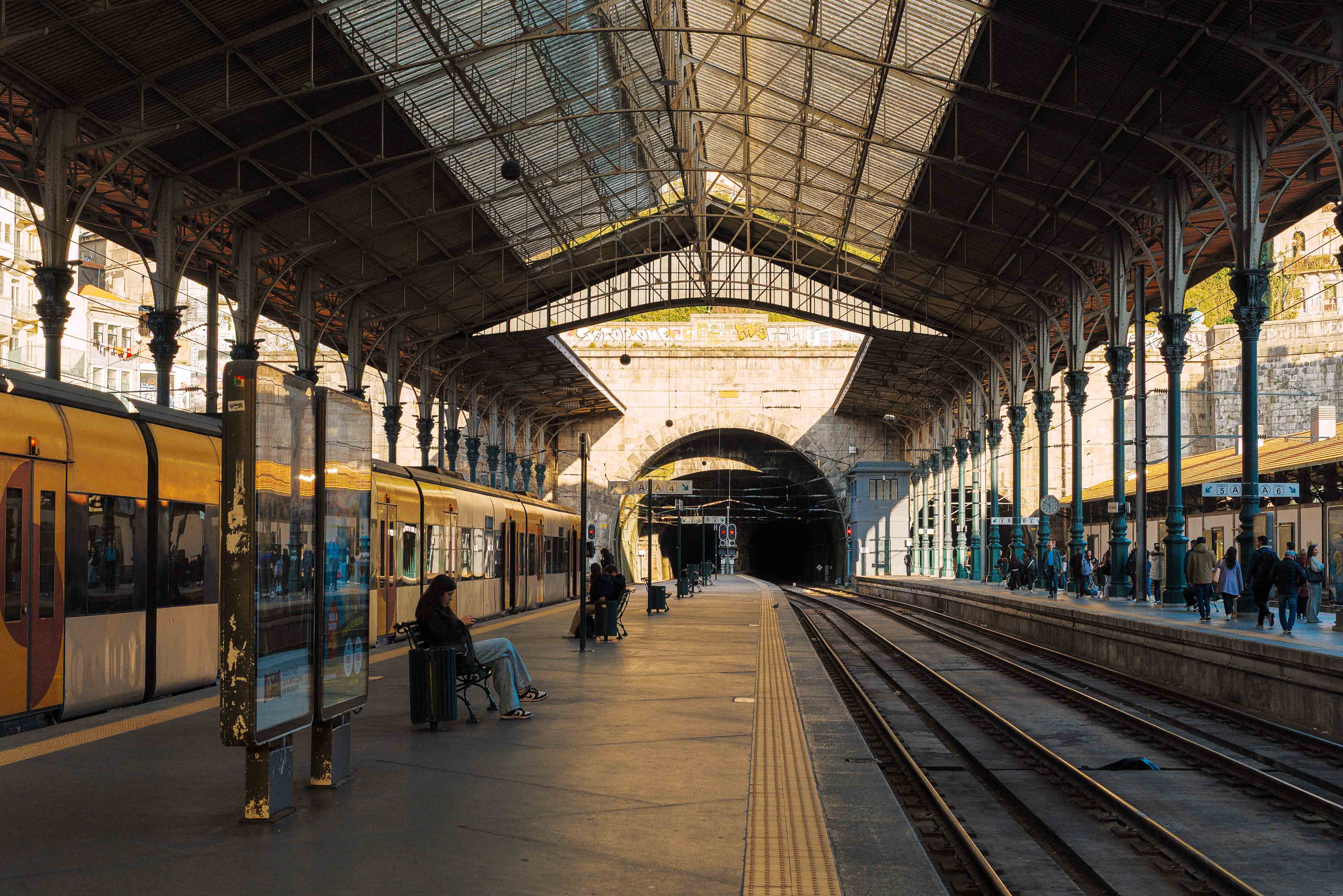 Interior view of São Bento railway station showing iron columns, a glass roof, trains waiting at the platforms, and passengers scattered along the concourse leading into a hillside tunnel.
