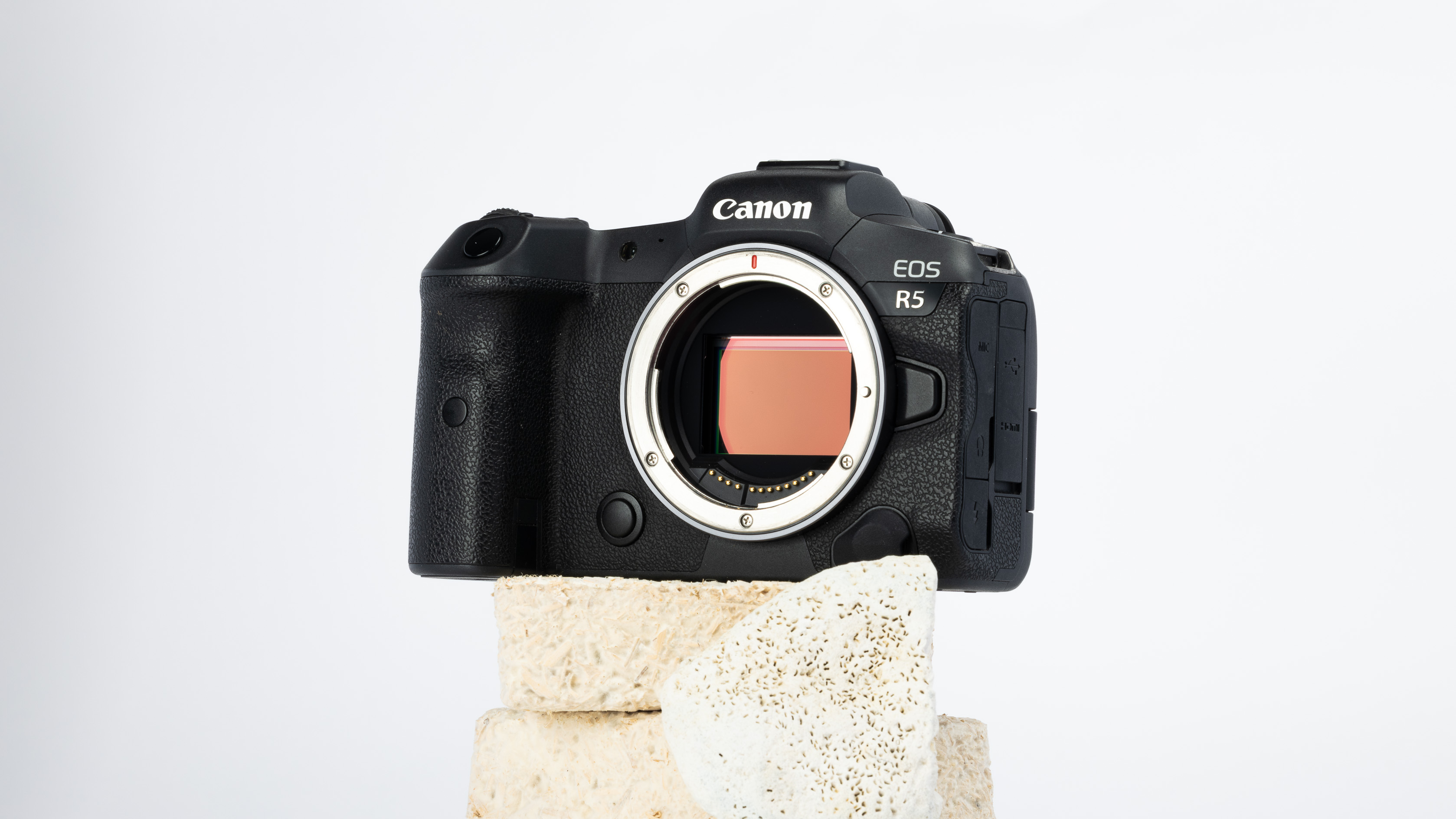 A product shot of a secondhand Canon EOS R5 camera propped up on a rocky podium on an off white background.
