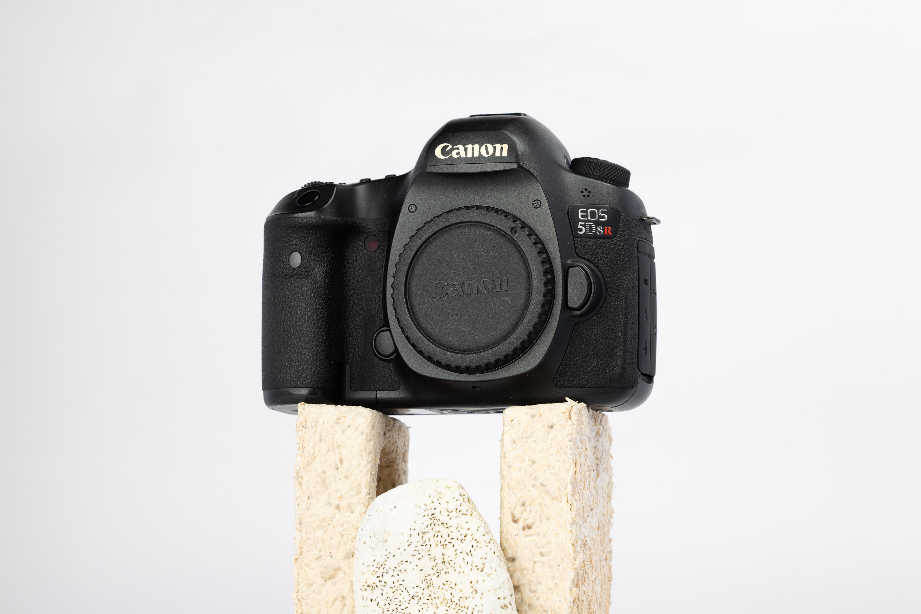 A product shot of a second hand Canon 5DS R camera propped up on a rocky podium on an off white background