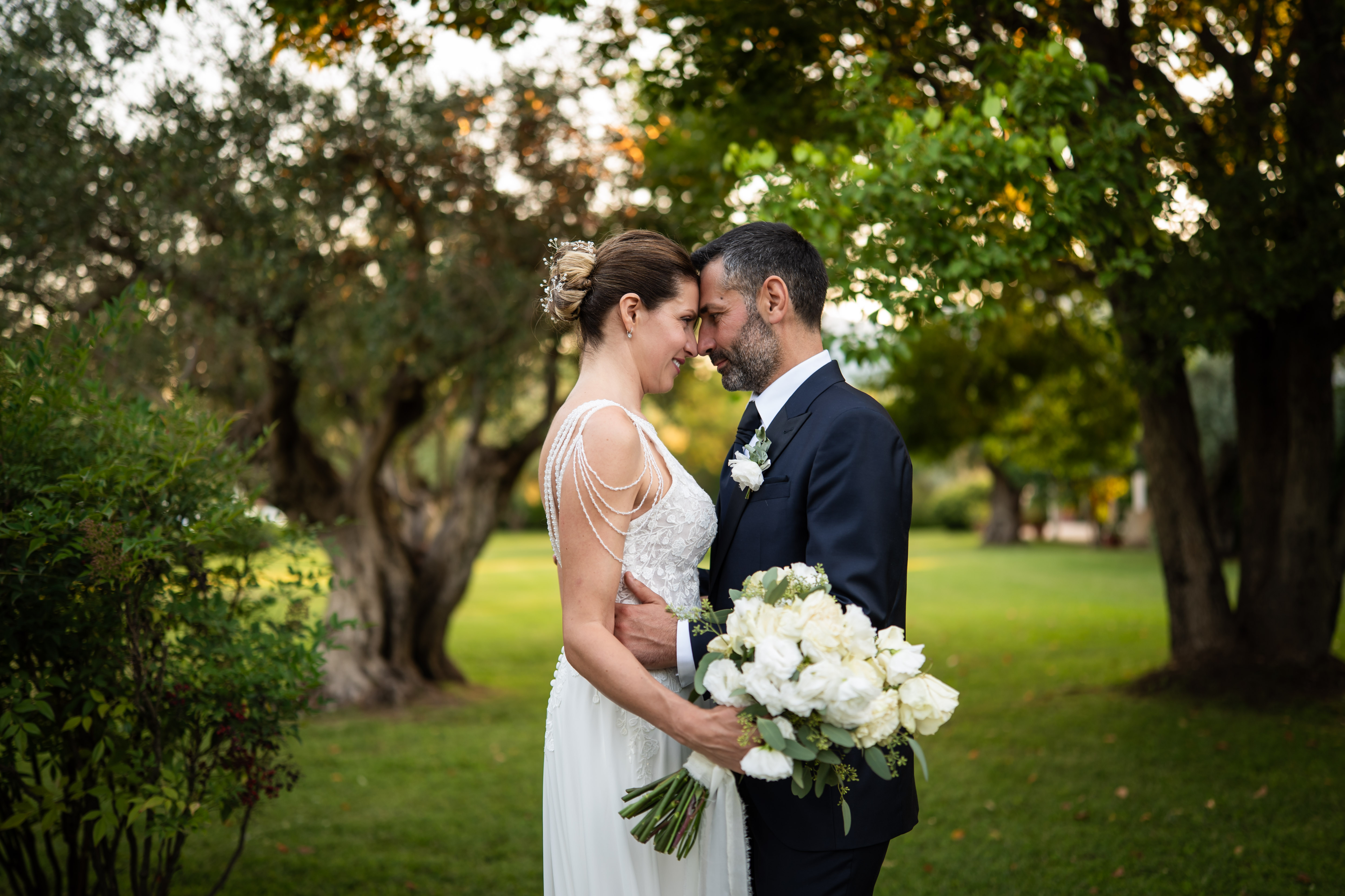 Sposi che si guardano fronte a fronte in un giardino verde, lei con bouquet di rose bianche, sfondo di alberi sfocato.