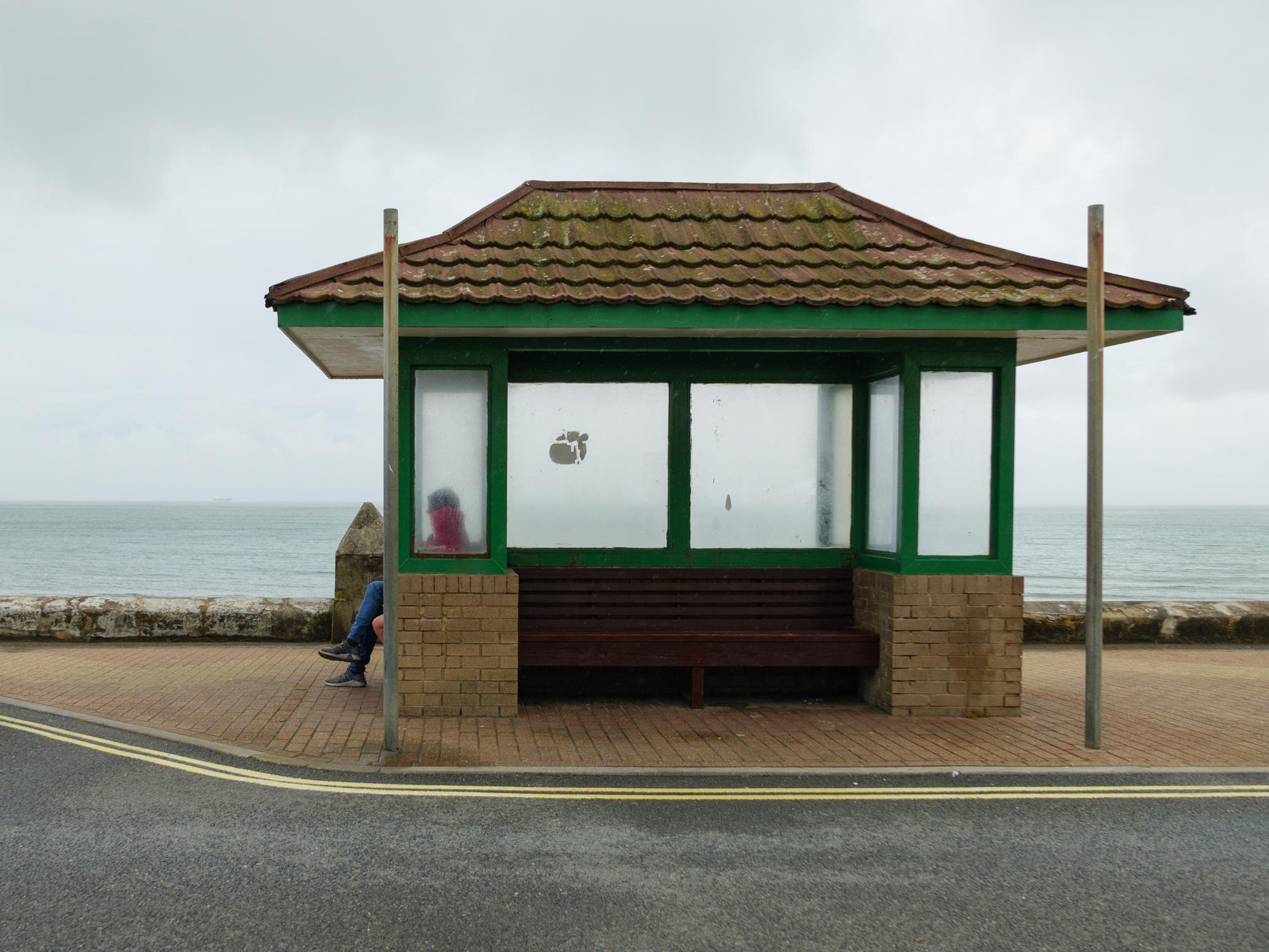 A shelter on the seafront keeps two people in waterproofs dry, with the sea in the background. Shot on the Panasonic Lumix DC-L10.