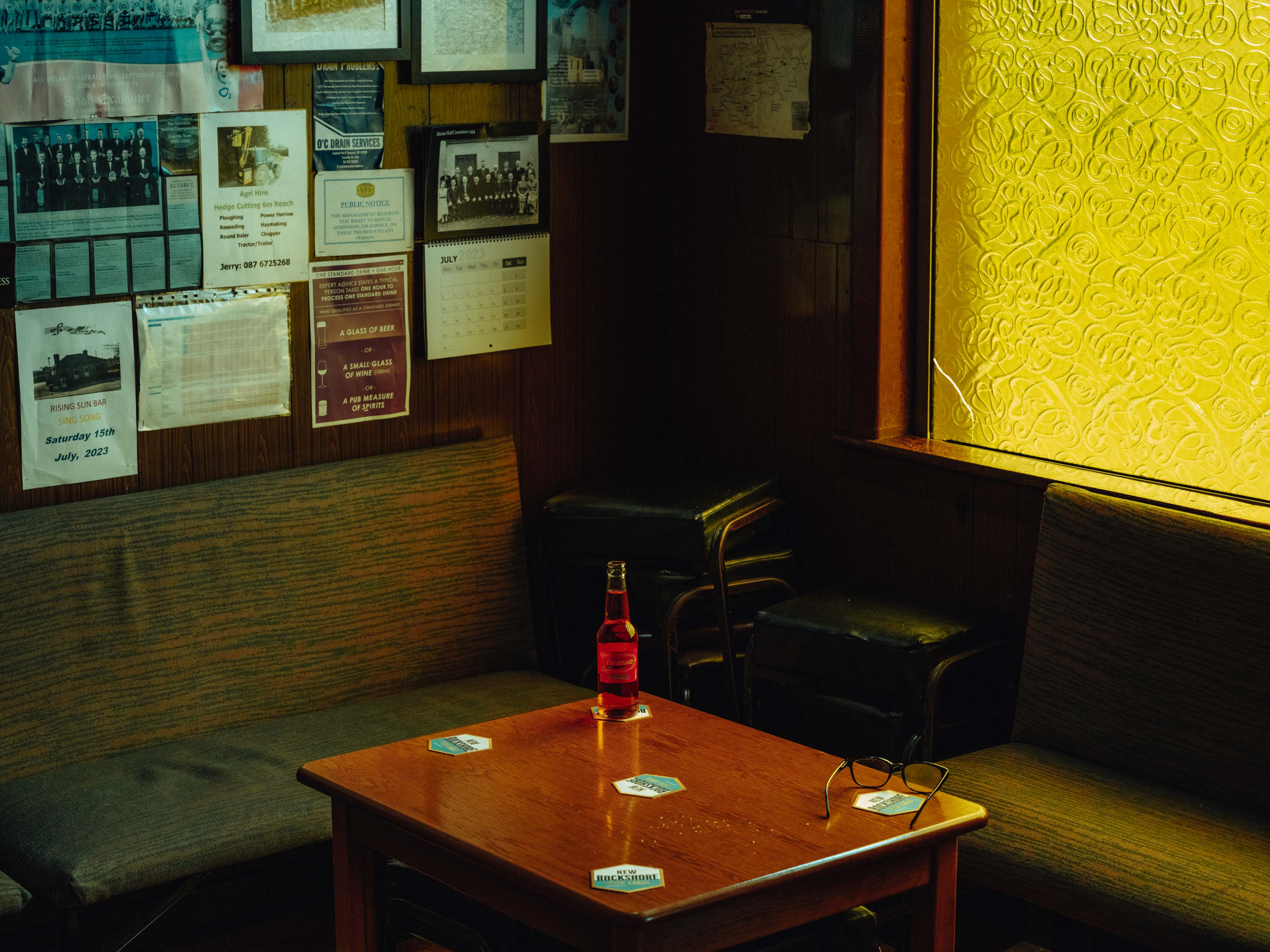 Photo d'une bouteille sur une table entourée de chaises vert foncé éclairées par une fenêtre translucide jaune, prise par Ian Howorth avec un Hasselblad X2D 100C.