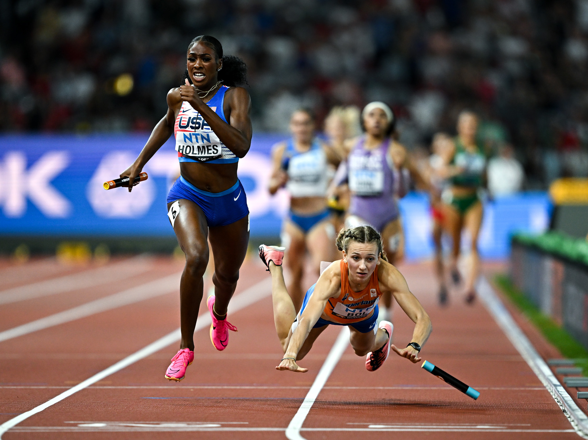An action shot of a relay race that shows one runner falling to the ground as the one beside them storms ahead. By Sam Barnes.