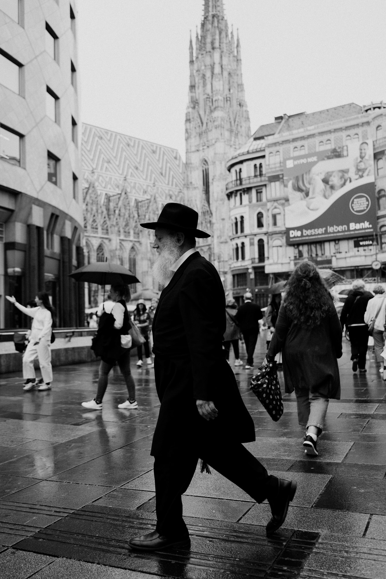 Schwarz-Weiß-Foto einer Straßenszene im Wiener Stadtzentrum. Im Vordergrund geht ein Mann mit weißem Vollbart und dunkler Kleidung über nasses Pflaster, während im Hintergrund der Stephansdom und weitere Passant:innen zu sehen sind. Aufgenommen von Dhan Limwattana mit einer Nikon Z5 II.