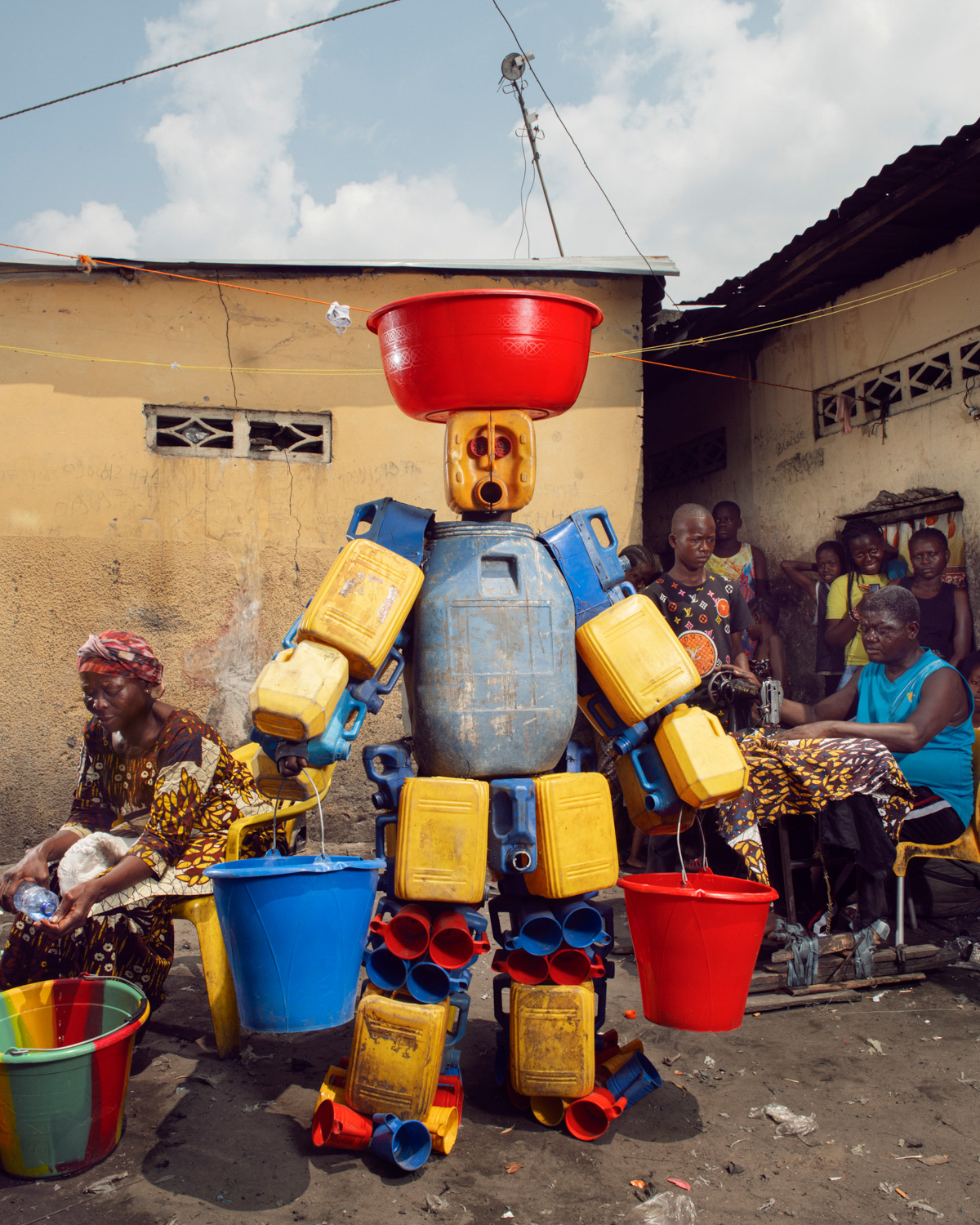 Eine Figur aus Kanistern und Eimern, die als Teil von Stephan Gladieus Kunstprojekt Homo Detritus über Abfall und Identität in Kinshasa zwischen Wäscherinnen steht.