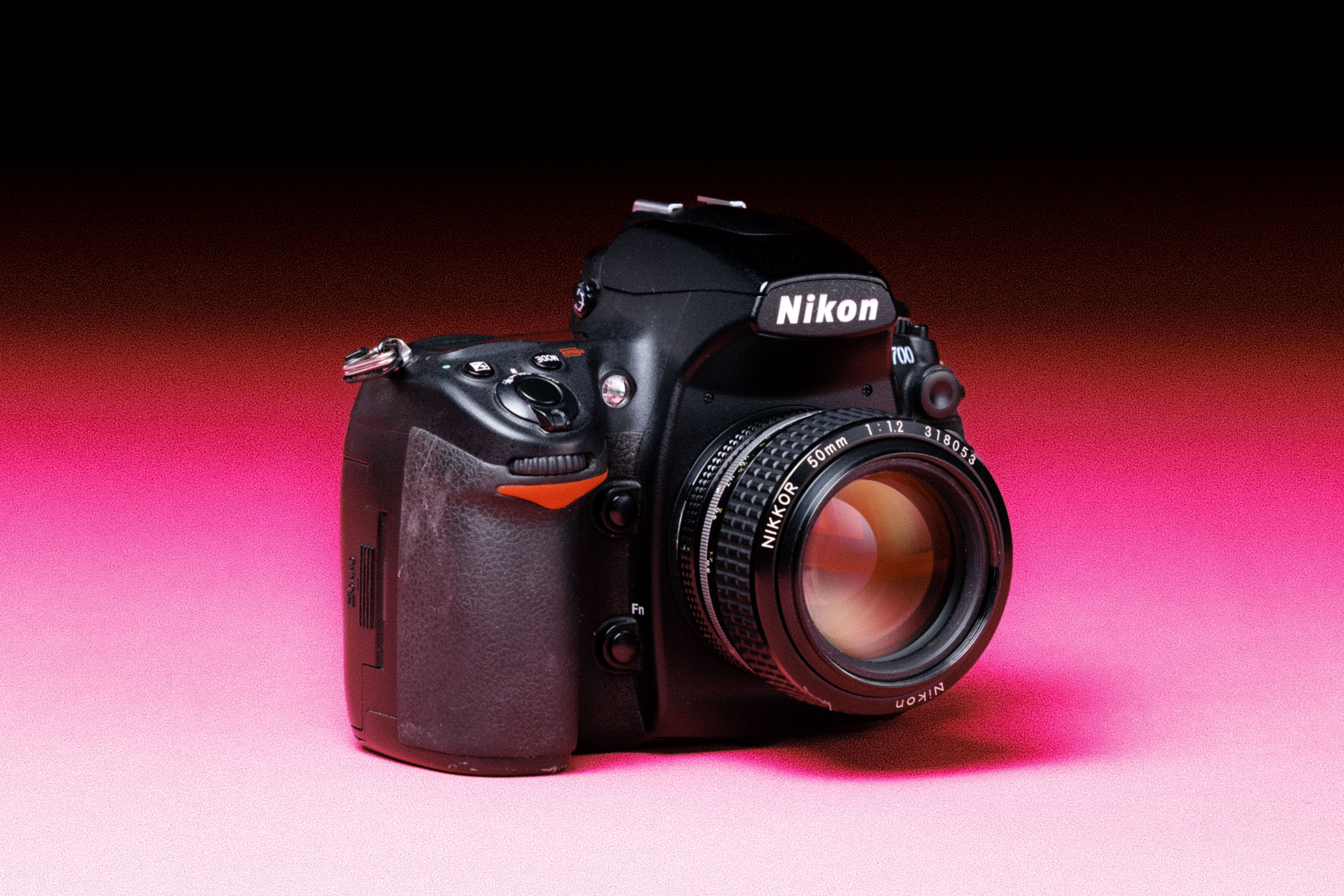 A Nikon D700 camera with a Nikon 50mm f/1.2 AIS lens, with an added film-like grain effect on a graduated pink background.
