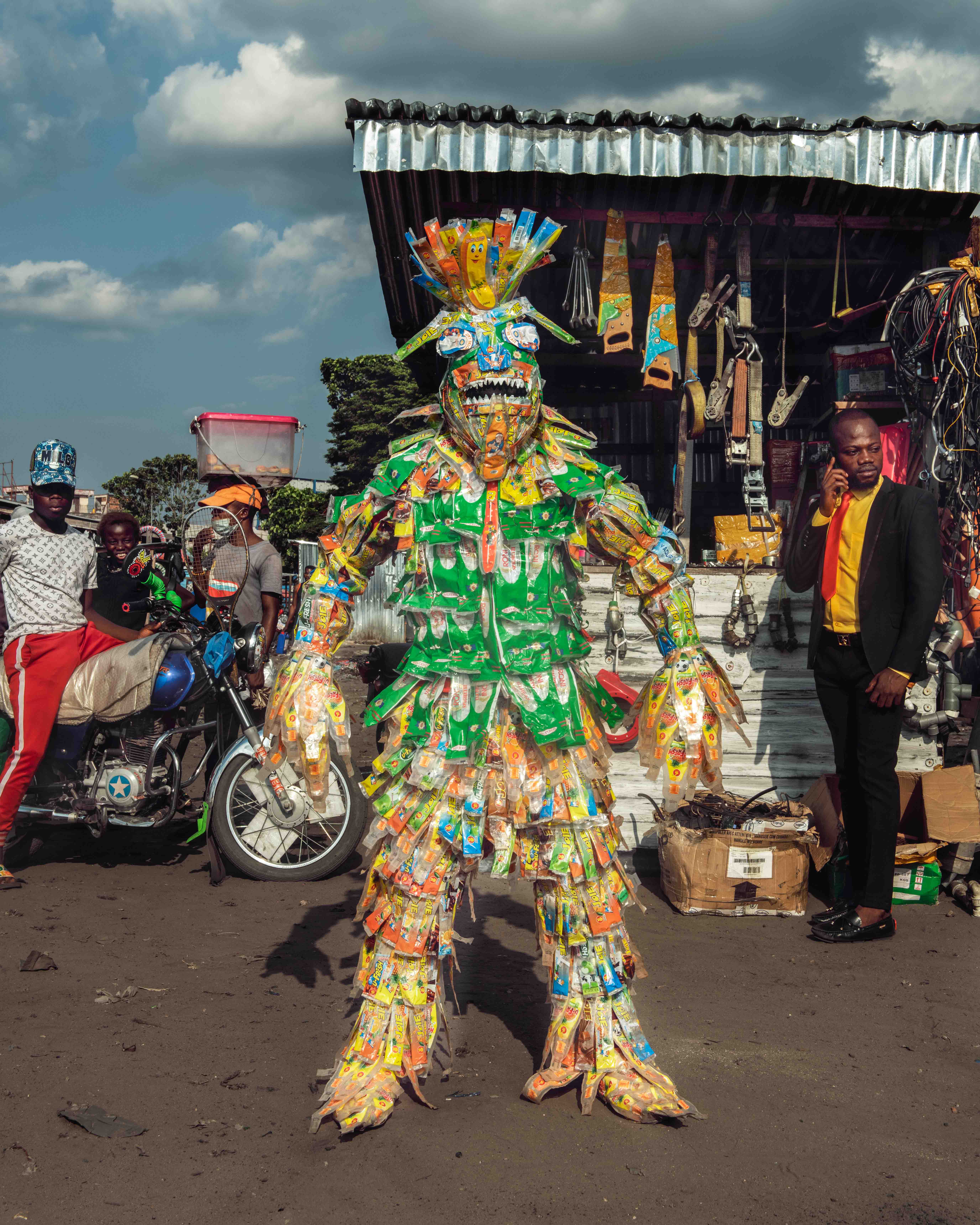 Eine Figur aus Stephan Gladieus Homo Detritus, die in Tütenabfall gehüllt ist und trotzig auf einem belebten kongolesischen Straßenmarkt posiert.