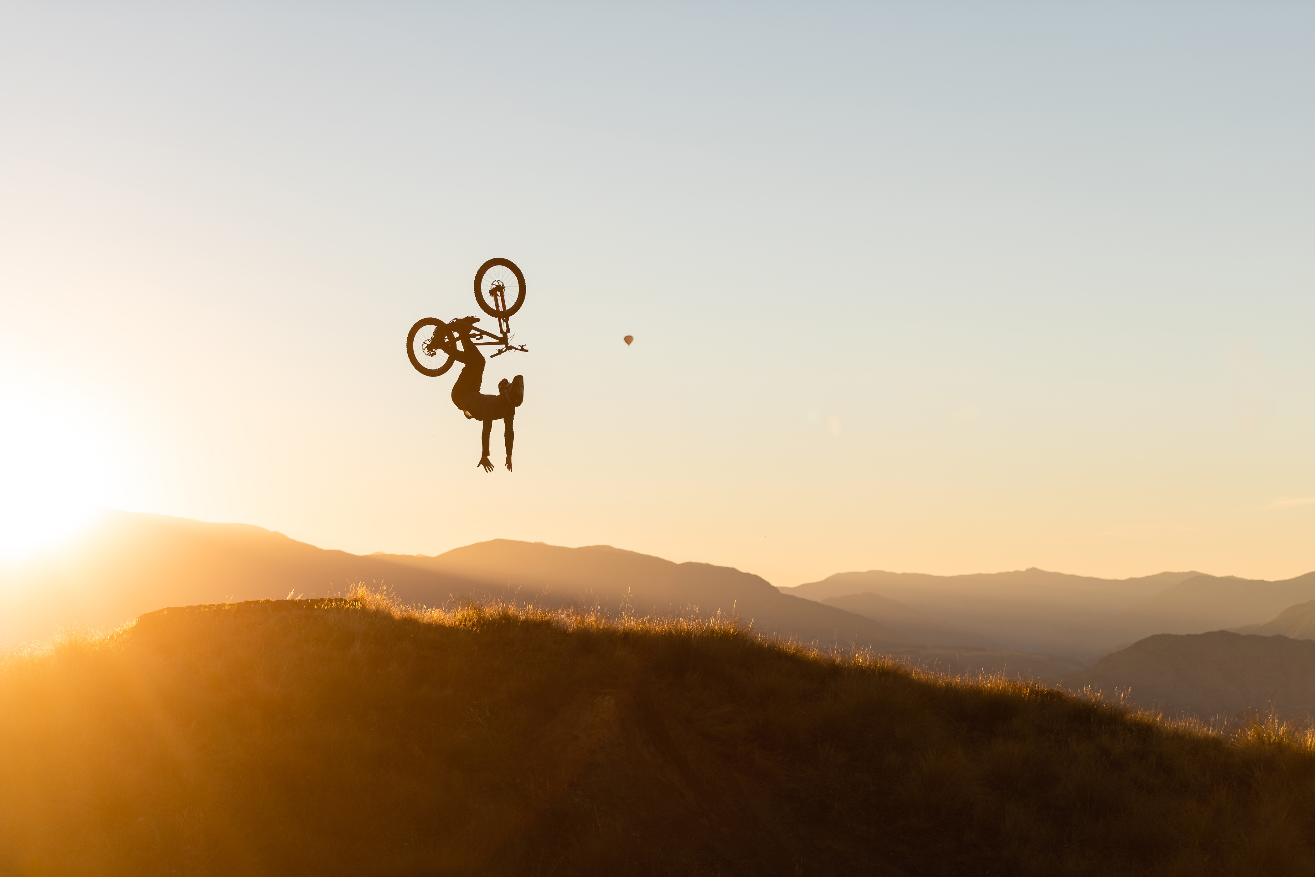 Silhouette of a cyclist doing a backflip at sunset over a mountain range.