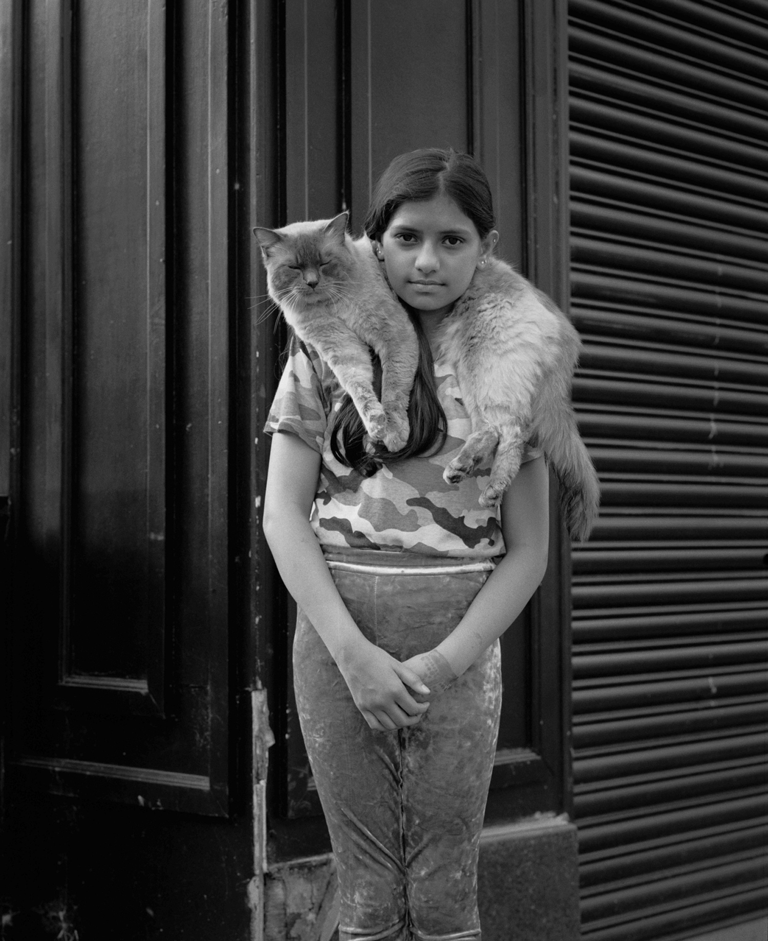 Black-and-white street portrait of a teenager with cat around their neck, standing in front of a dark building with the shop shutters down, photo by Simon Murphy