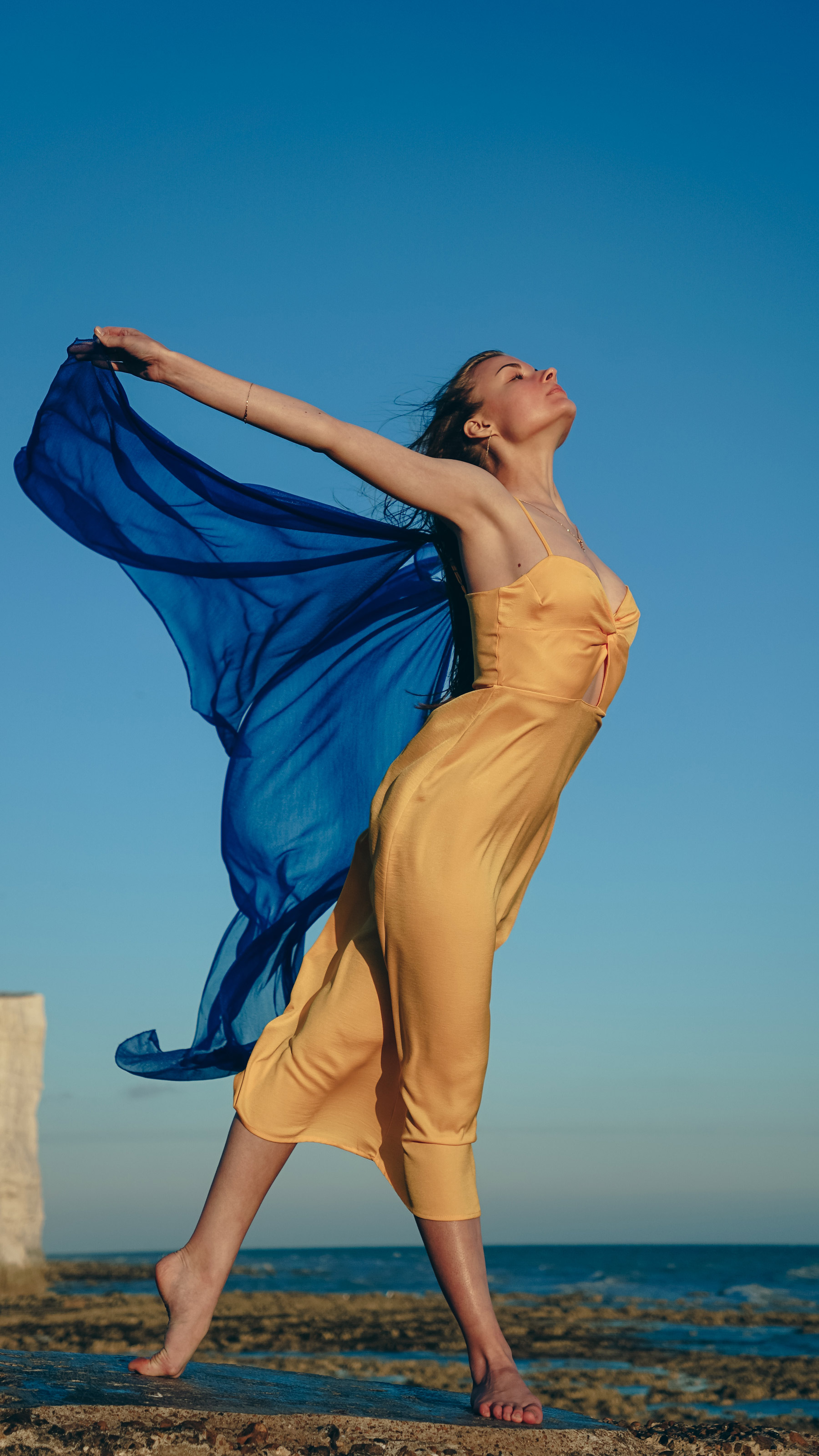 A model wearing the Ukraine colours — a yellow dress and a blue scarf — poses on a beach under a bright blue sky, shot by Nataliia Petrishyna with a Sony A6000