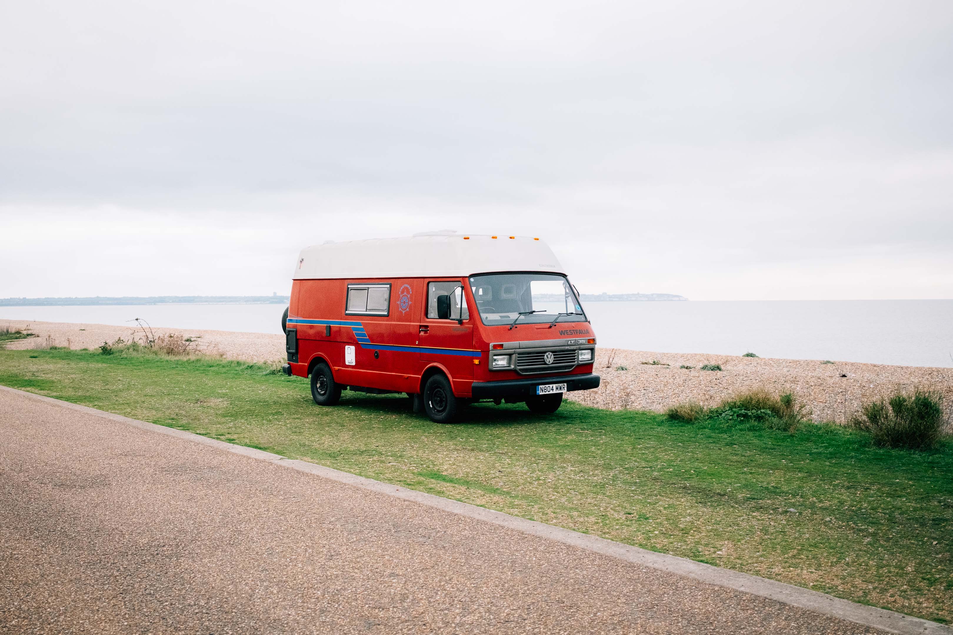 A vintage red Volkswagen camper parked by a shingle beach in Kent. Taken on the Fujifilm X100VI by Connor Redmond.